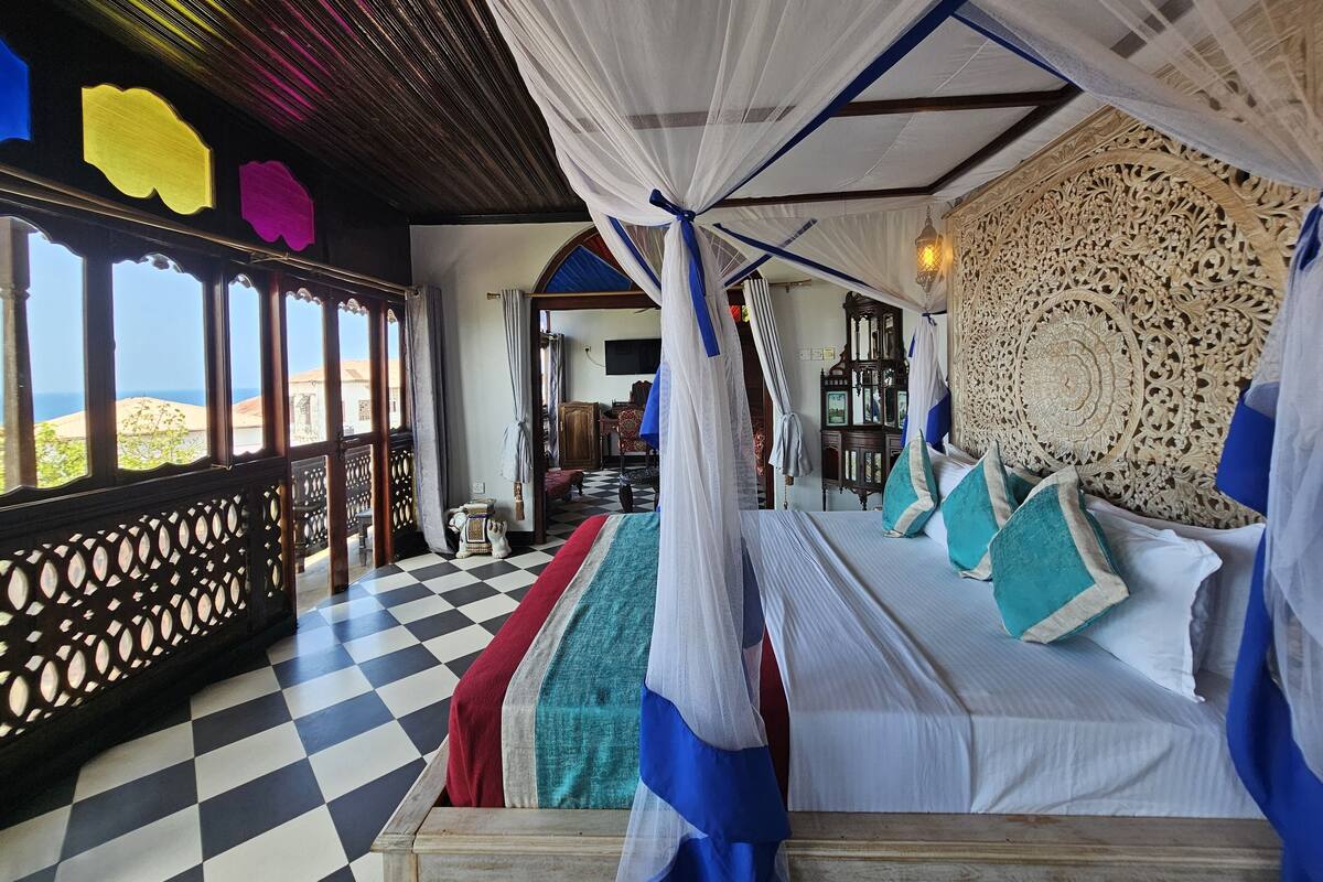 A room with a checkered floor, a bed with white and blue bedding, a canopy bed, and a view of the sea.