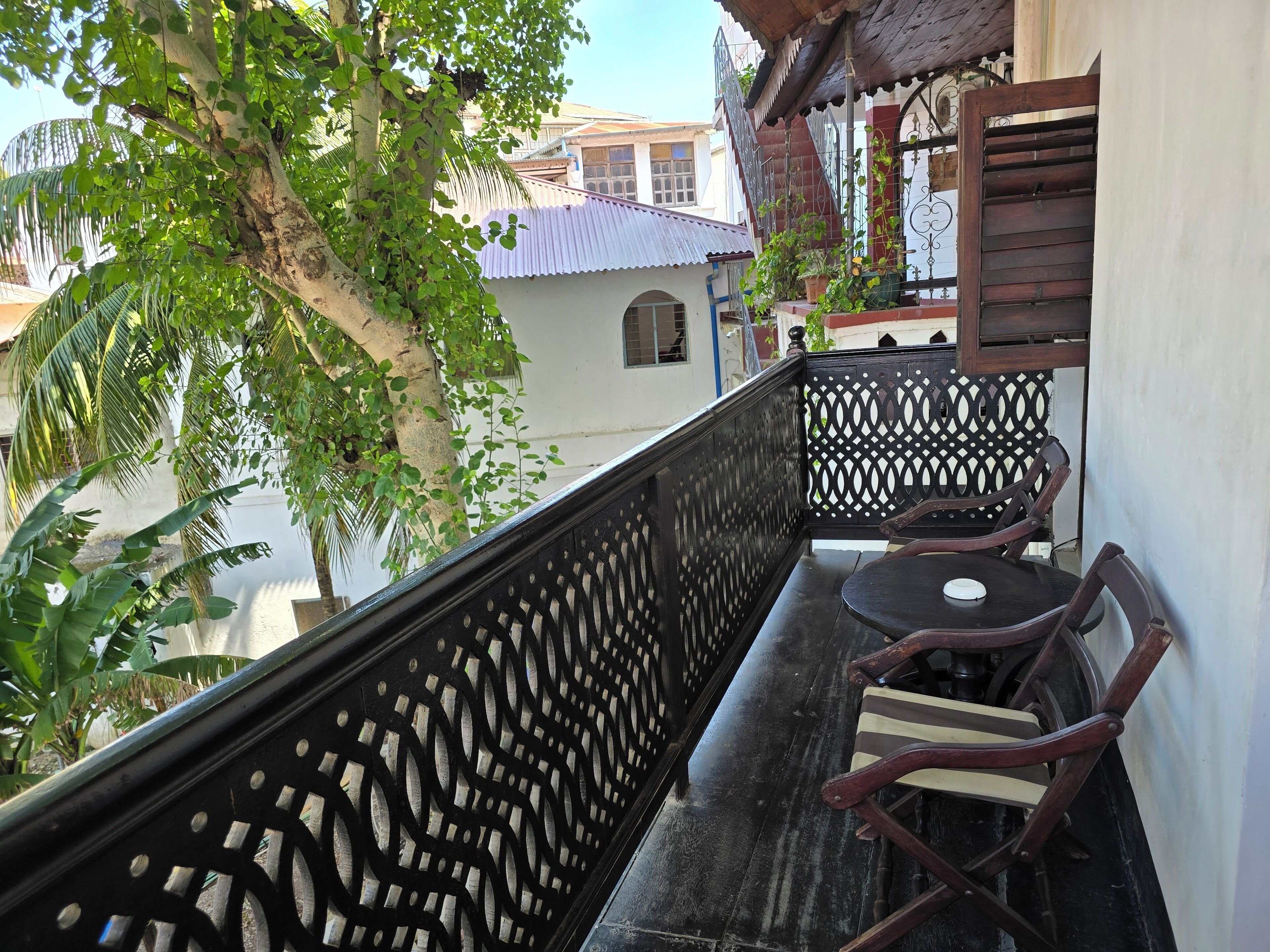Un balcon avec une table et des chaises en bois, une balustrade à motifs et une vue sur un bâtiment dont le toit est fait de tôles ondulées.