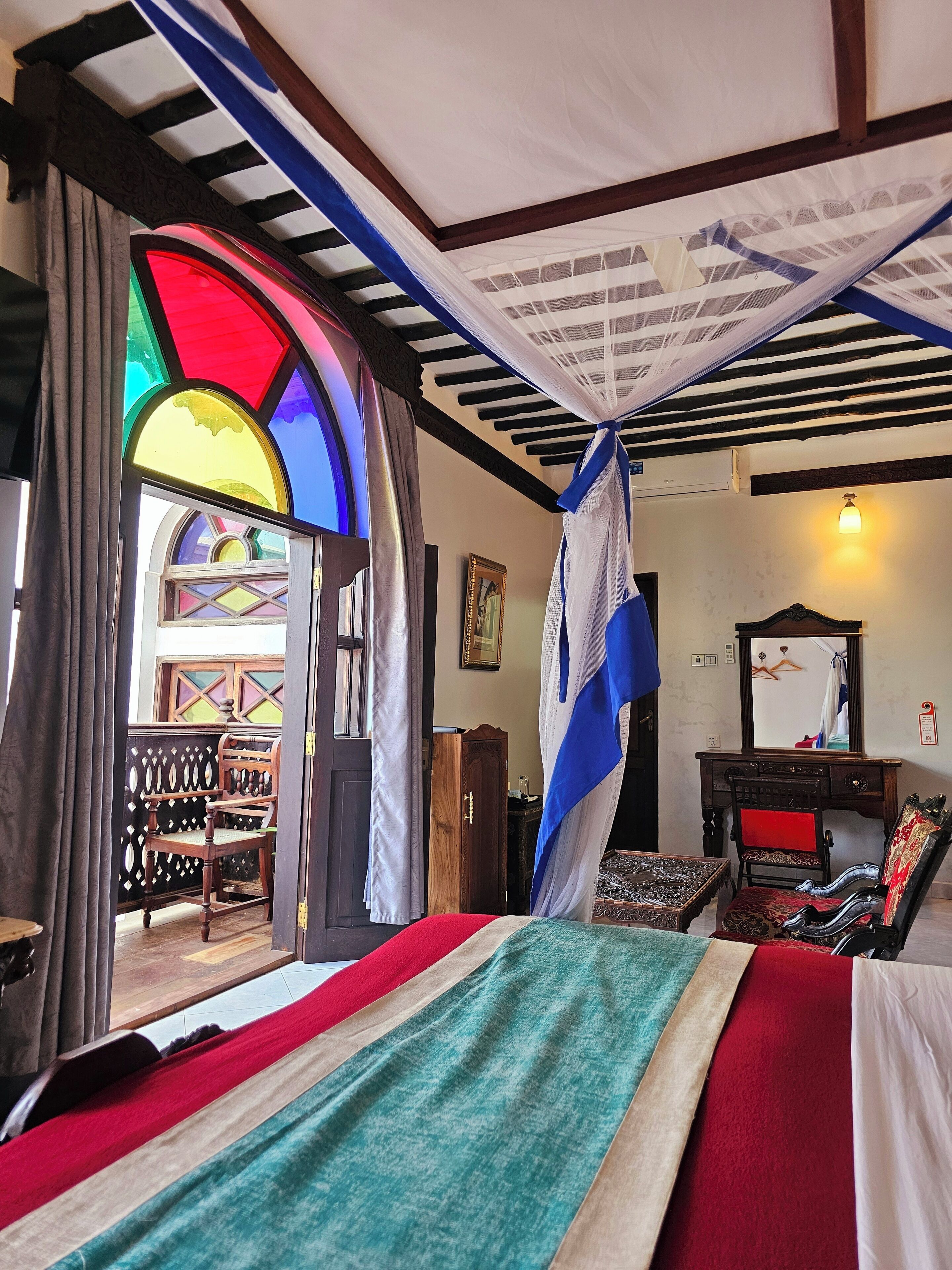 A room with a bed, a wooden desk, a chair, and a window with stained glass.