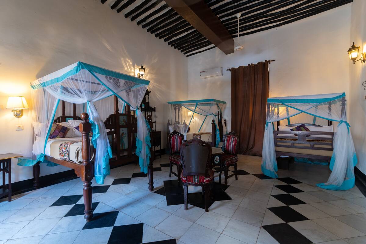 A room with a bed under a canopy, a chair, a table, and a wall-mounted lamp.