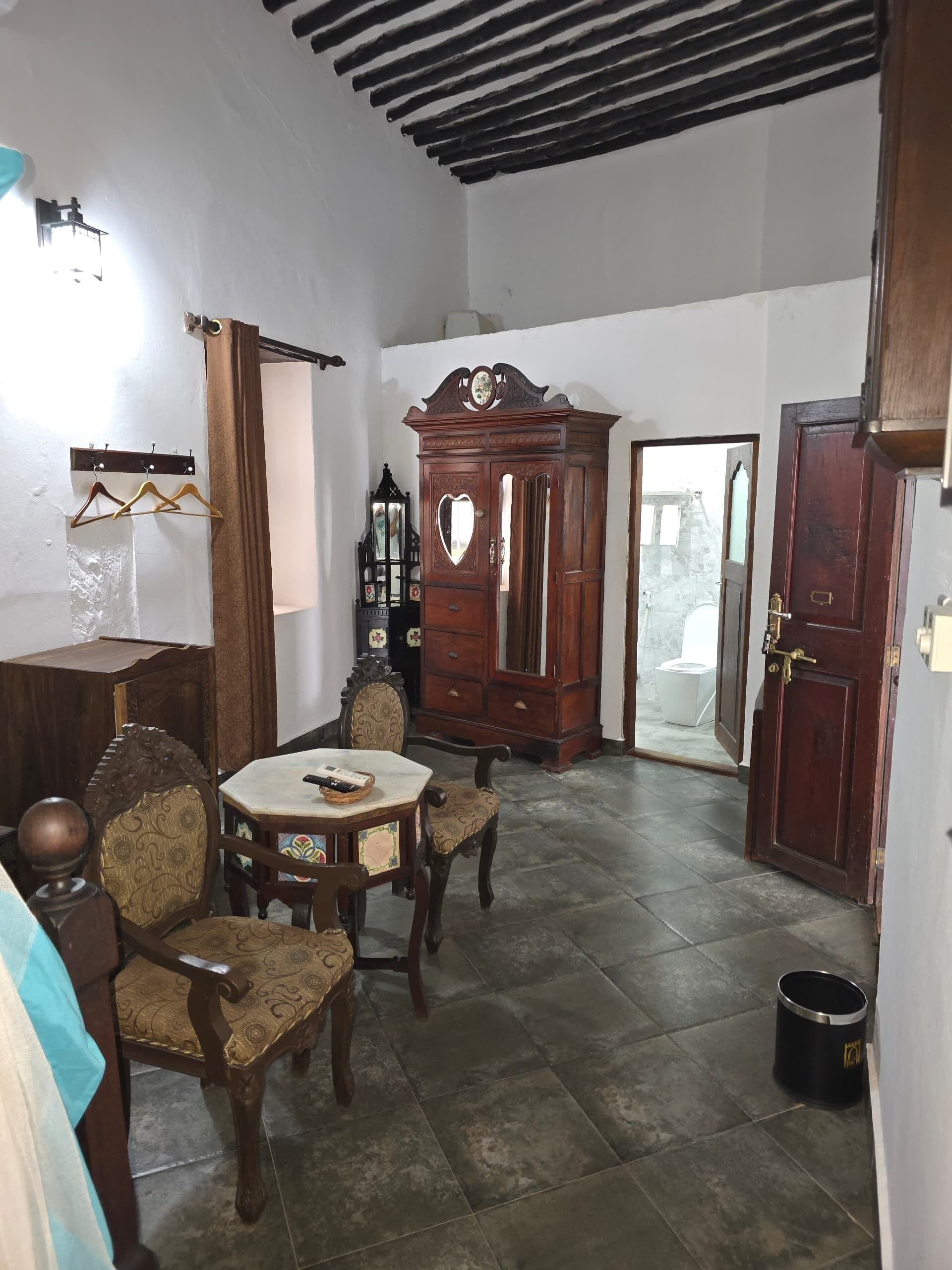 A dining area with a round table and four chairs, a wooden cabinet with a heart-shaped mirror, and a coat rack with hangers.