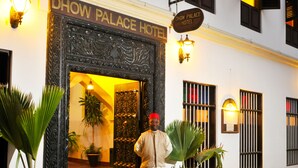 The exterior of Dhow Palace Hotel with a traditional architectural style, featuring a prominent entrance, potted plants, and a signboard.
