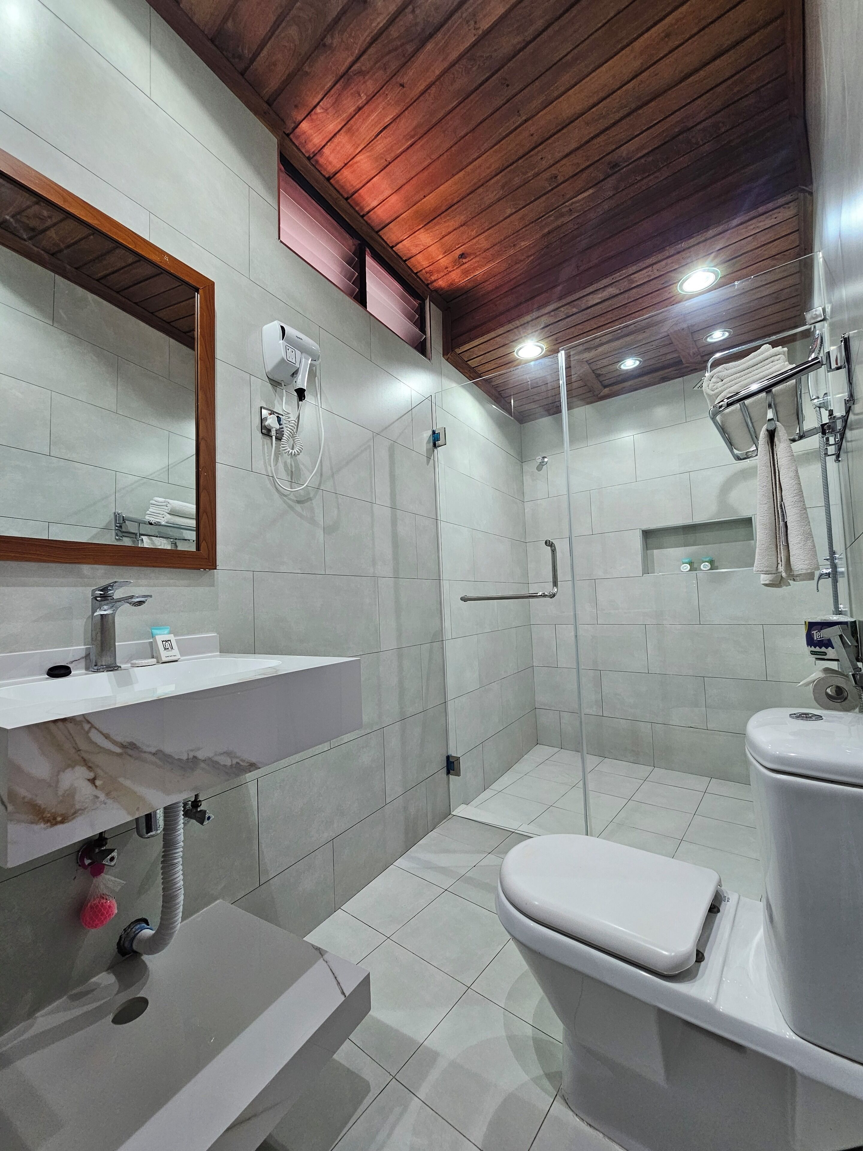 Un baño moderno con lavamanos de mármol, inodoro, ducha con puerta de vidrio y techo de madera.
