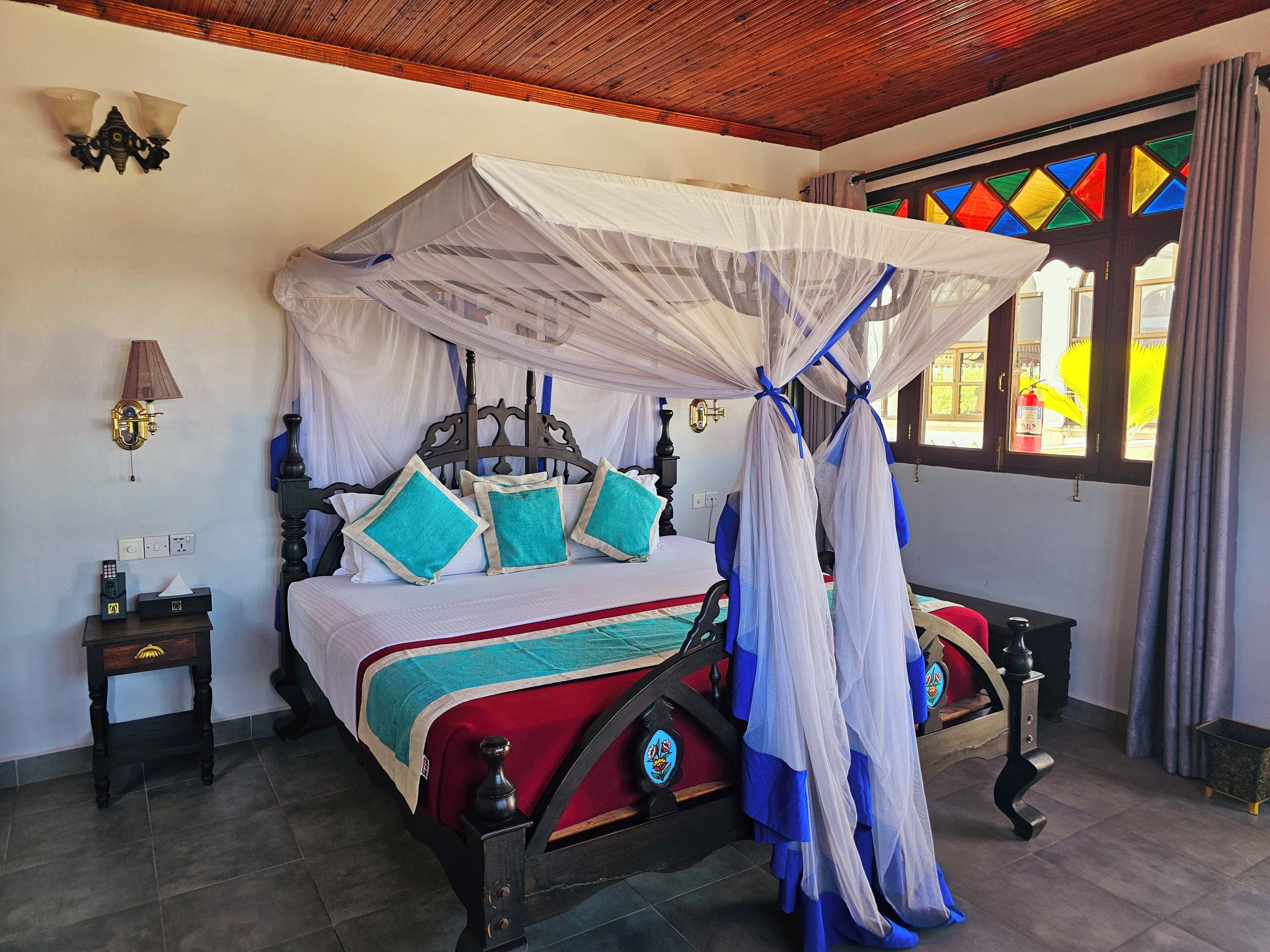 A bed with a canopy, a nightstand, a lamp, and a window with colorful stained glass.