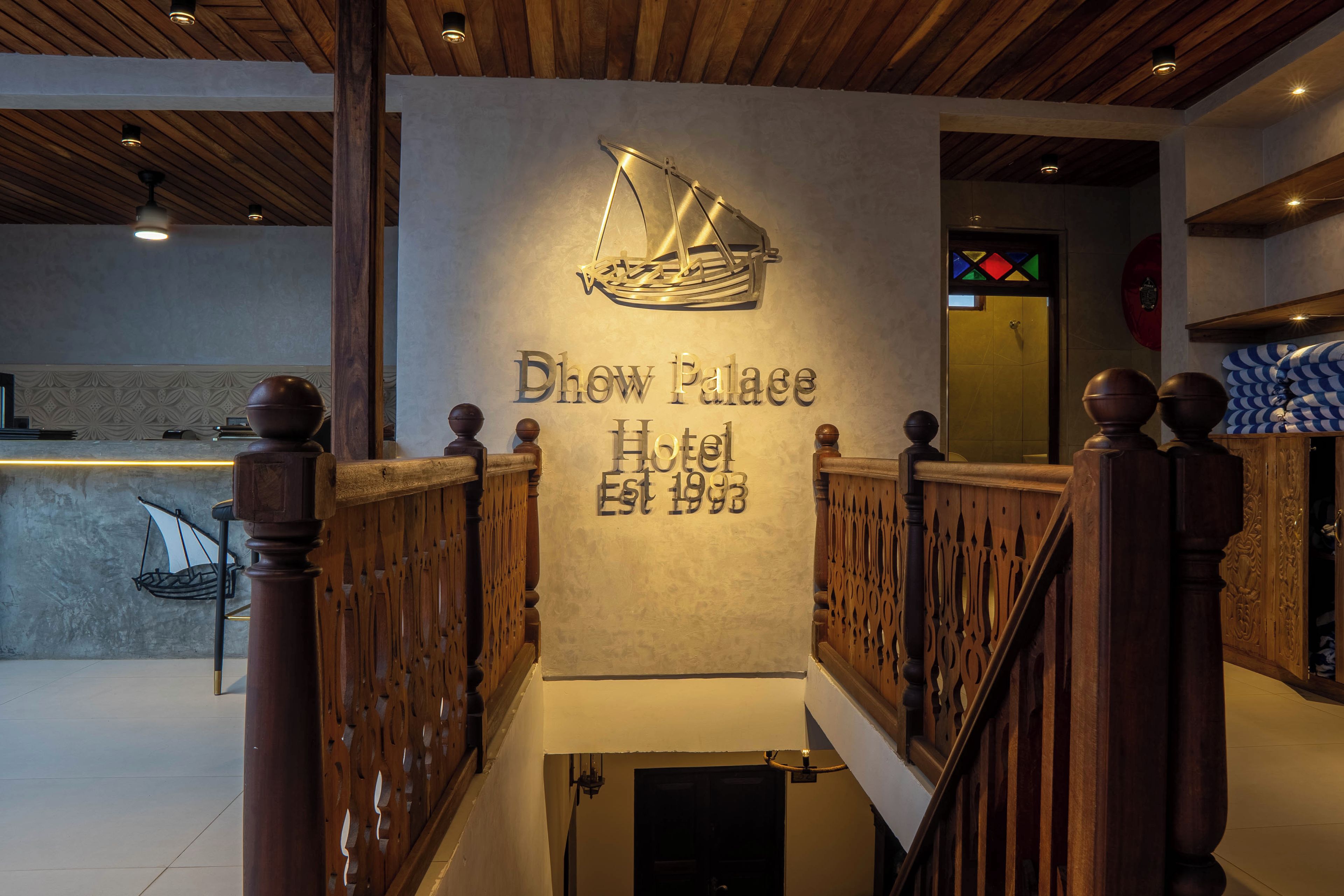 A reception area with a wooden staircase, a wall-mounted sign for 'Dhow Palace Hotel Est 1993', and a decorative ship emblem.