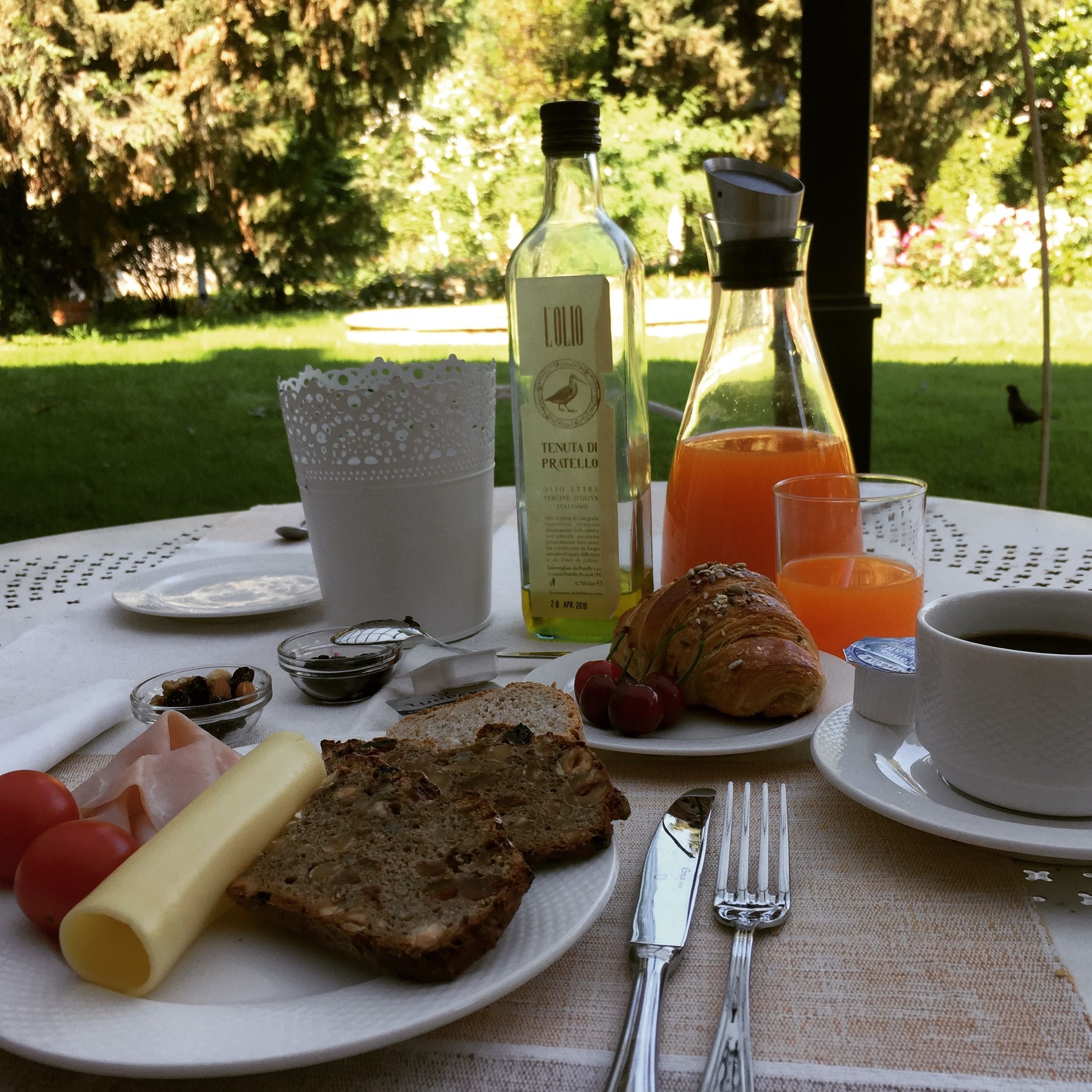 Une table dressée pour le petit-déjeuner, avec des croissants, du pain, du fromage et des fruits, et une vue sur un jardin.