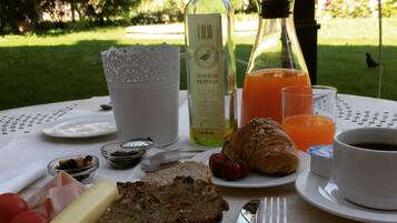Une table dressée pour le petit-déjeuner, avec des croissants, du pain, du fromage et des fruits, et une vue sur un jardin.