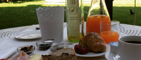 A breakfast setting with croissants, bread, cheese, and fruit on a table with a view of a garden.