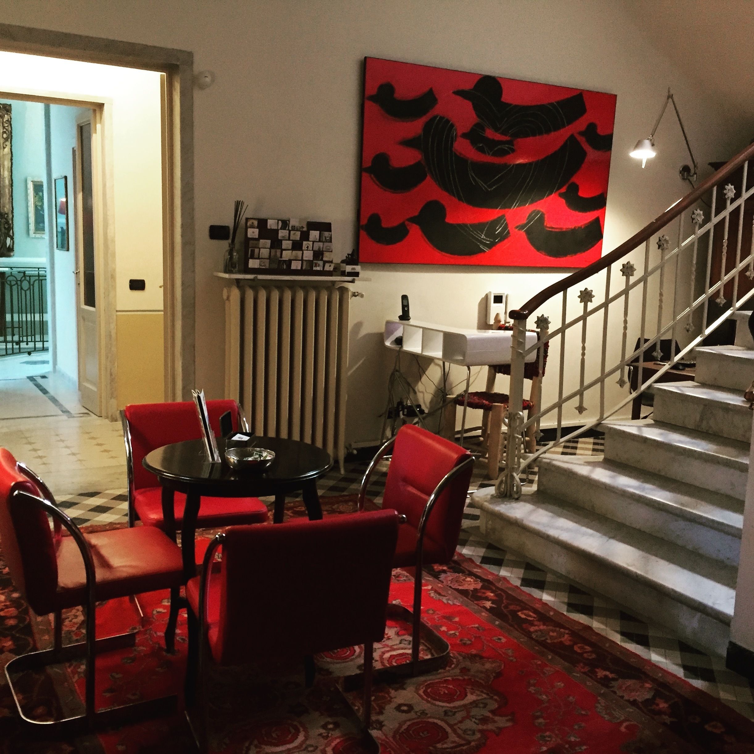 Un comedor con alfombra roja, una mesa negra y sillas rojas. Se observa una escalera con barandilla blanca y una pared roja con un cuadro abstracto en negro.