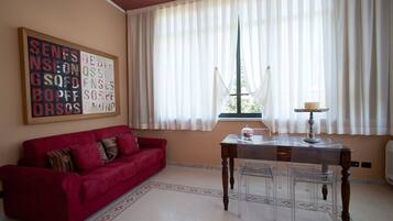 Una sala de estar con un sofá rojo, una mesa de madera con una lámpara y una ventana con cortinas de gasa.
