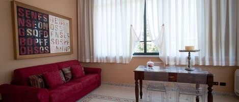 A living room with a red sofa, a wooden table with a lamp, and a window with sheer curtains.