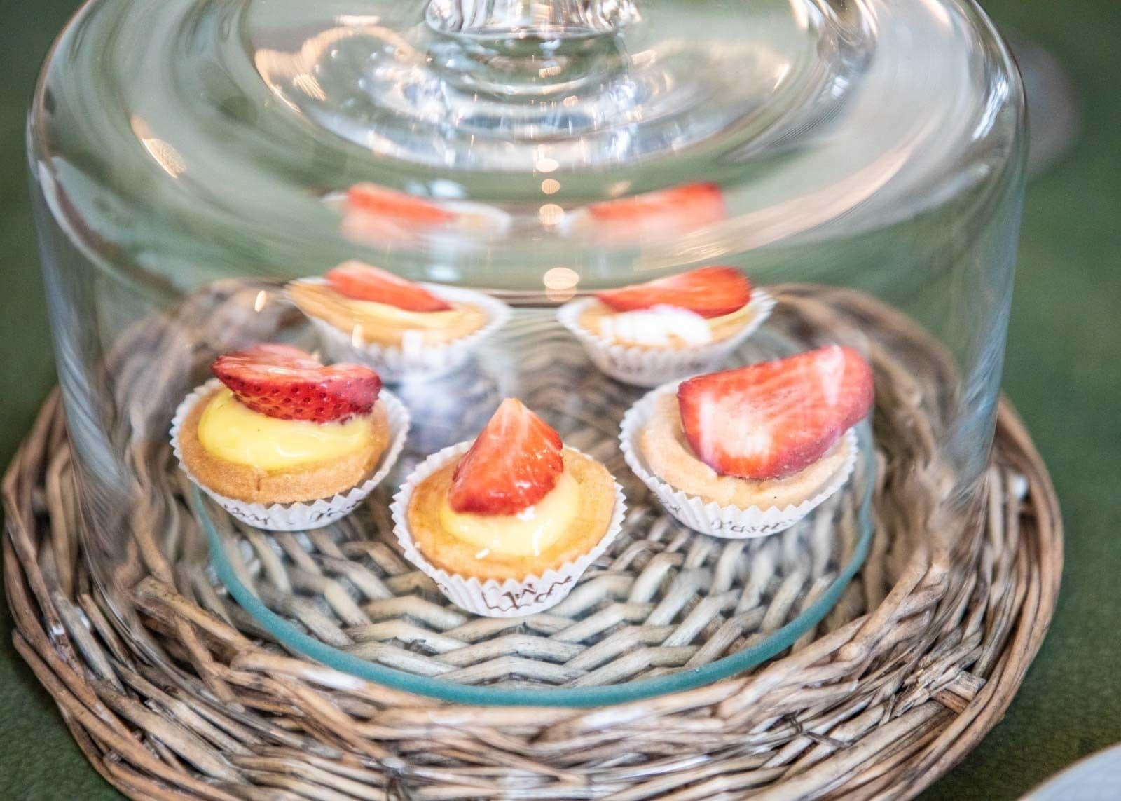 Eine Anordnung von Mini-Cheesecakes mit Erdbeeren unter einer Glaskuppel, platziert auf einer gewebten Matte.