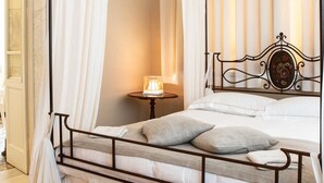 A four-poster bed with a metal frame, white linens, and grey pillows. There is a small round table with a lamp beside the bed. A wooden cabinet and a chair are visible in the background. - Villa Belverde (Carrara)