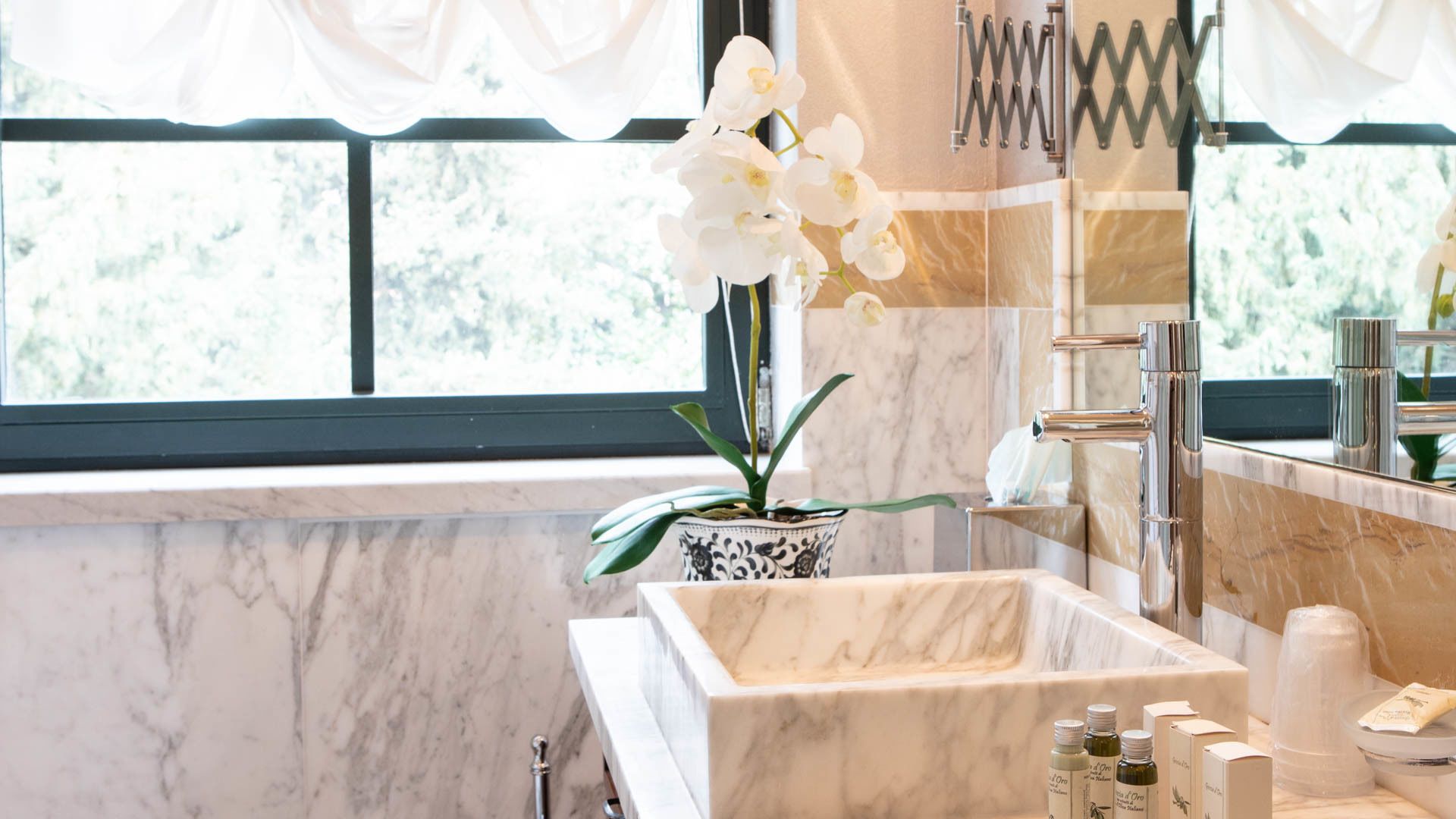 Une salle de bain avec un lavabo en marbre, une orchidée en pot, un grand miroir et une fenêtre avec des rideaux.
