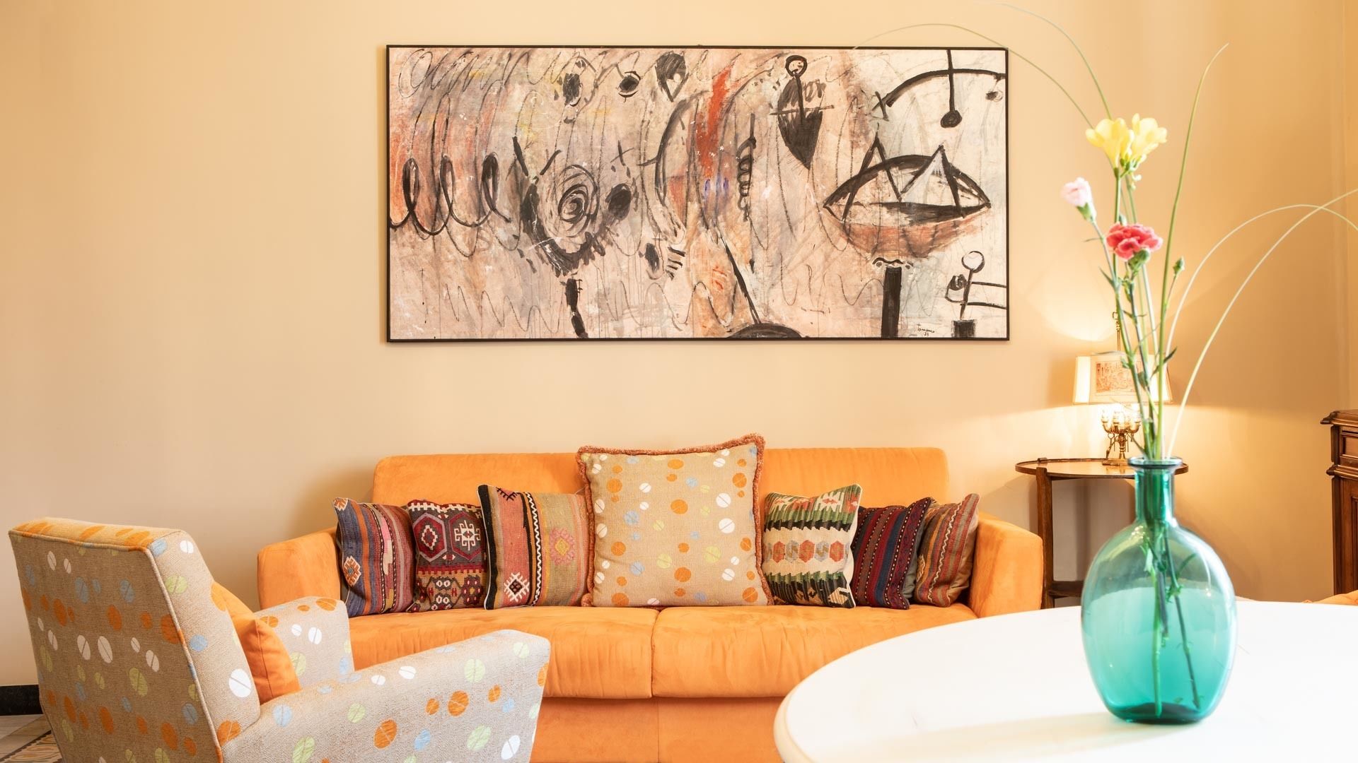 Un soggiorno con un divano, cuscini decorati, un vaso blu con fiori e un grande quadro astratto appeso al muro.