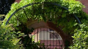 A covered outdoor seating area with two white chairs and a table, surrounded by lush greenery and a brick wall. - Villa Belverde (Carrara)