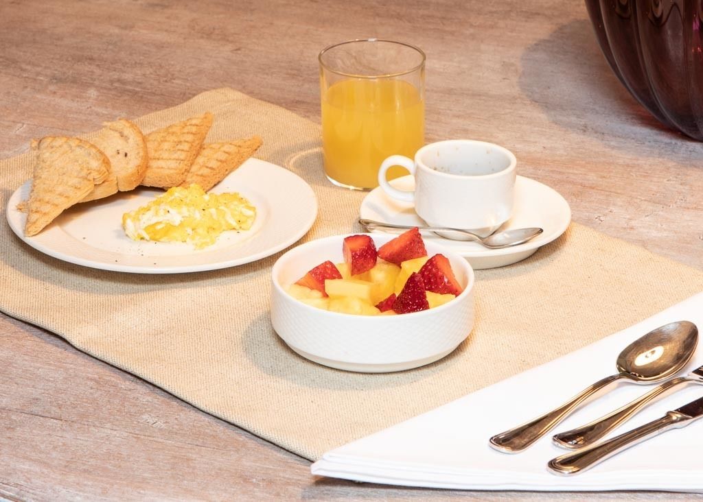 Una mesa de madera con waffles, huevos revueltos, un bol de fruta, un vaso de zumo de naranja y una taza de café.