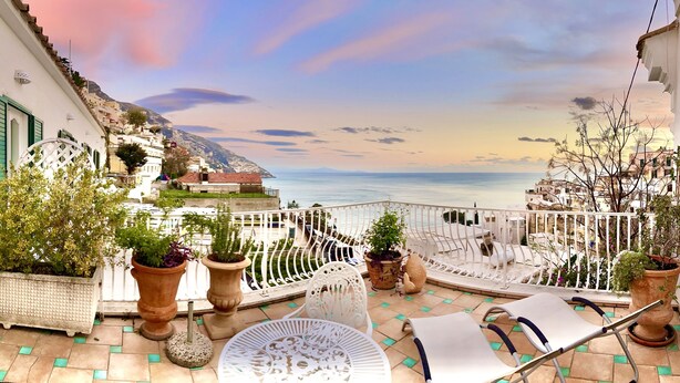 A balcony with potted plants, outdoor furniture, and a view of the sea and a mountainous landscape. - Hotel Villa delle Palme (Positano)