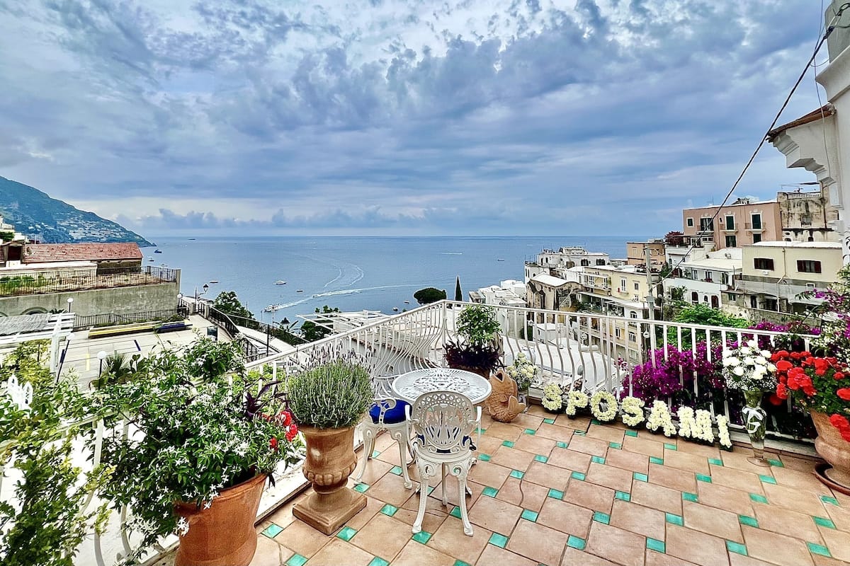 Un balcon agrémenté de plantes en pot et de mobilier d’extérieur, donnant sur une ville côtière avec ses bâtiments et la mer.