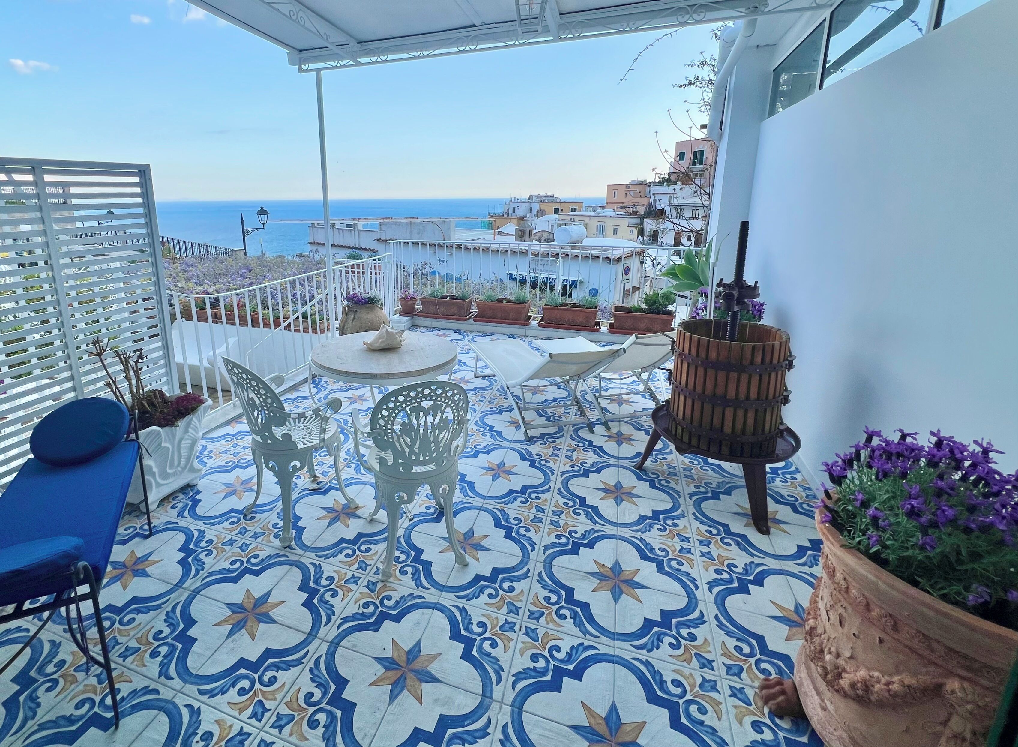 Une terrasse agrémentée de carreaux décoratifs, de meubles d’extérieur, de plantes en pot et offrant une vue sur la mer et les bâtiments.