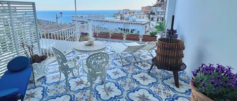 Une terrasse agrémentée de carreaux décoratifs, de meubles d’extérieur, de plantes en pot et offrant une vue sur la mer et les bâtiments.