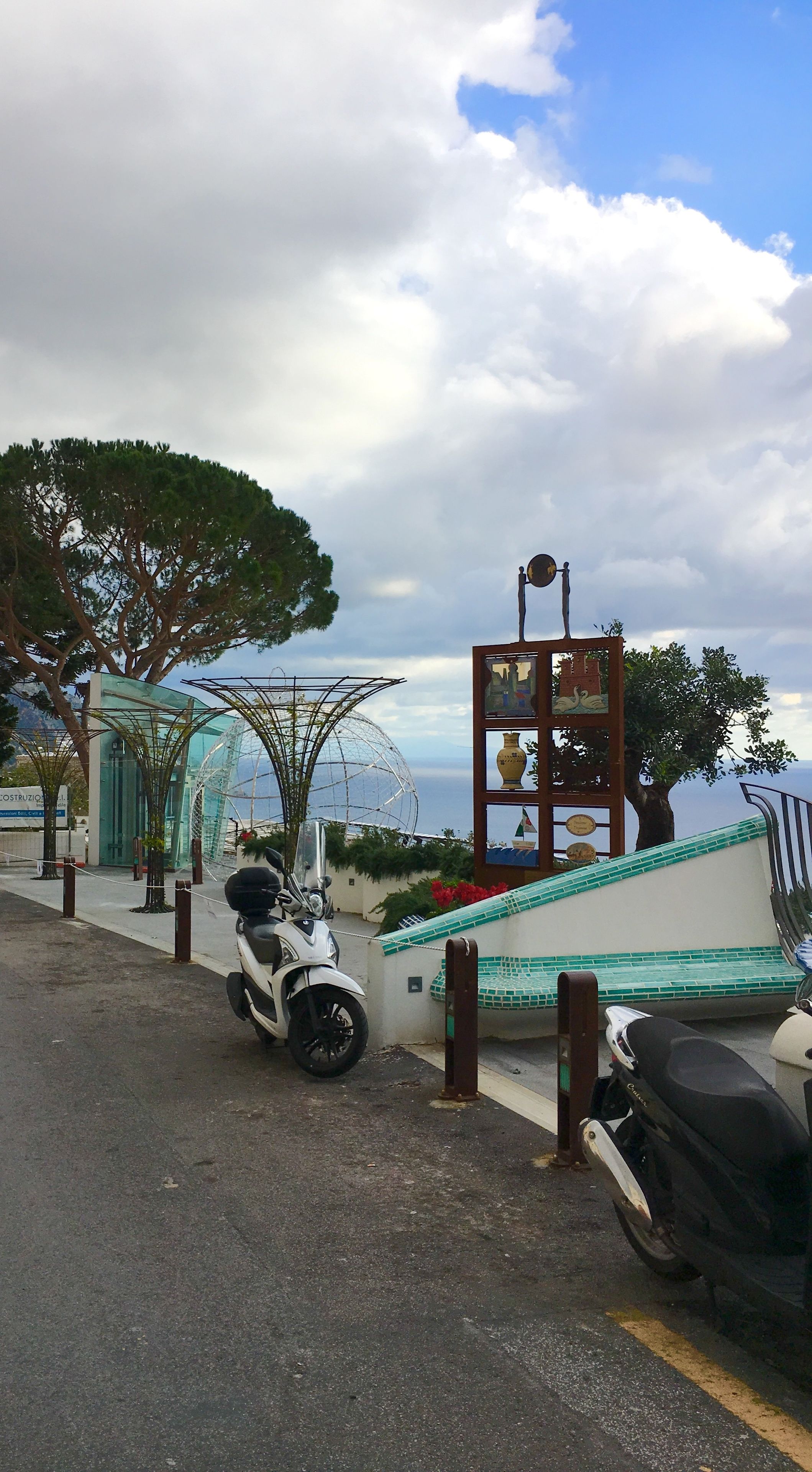 A parking area with scooters, a glass structure, and a decorative sign with various items.