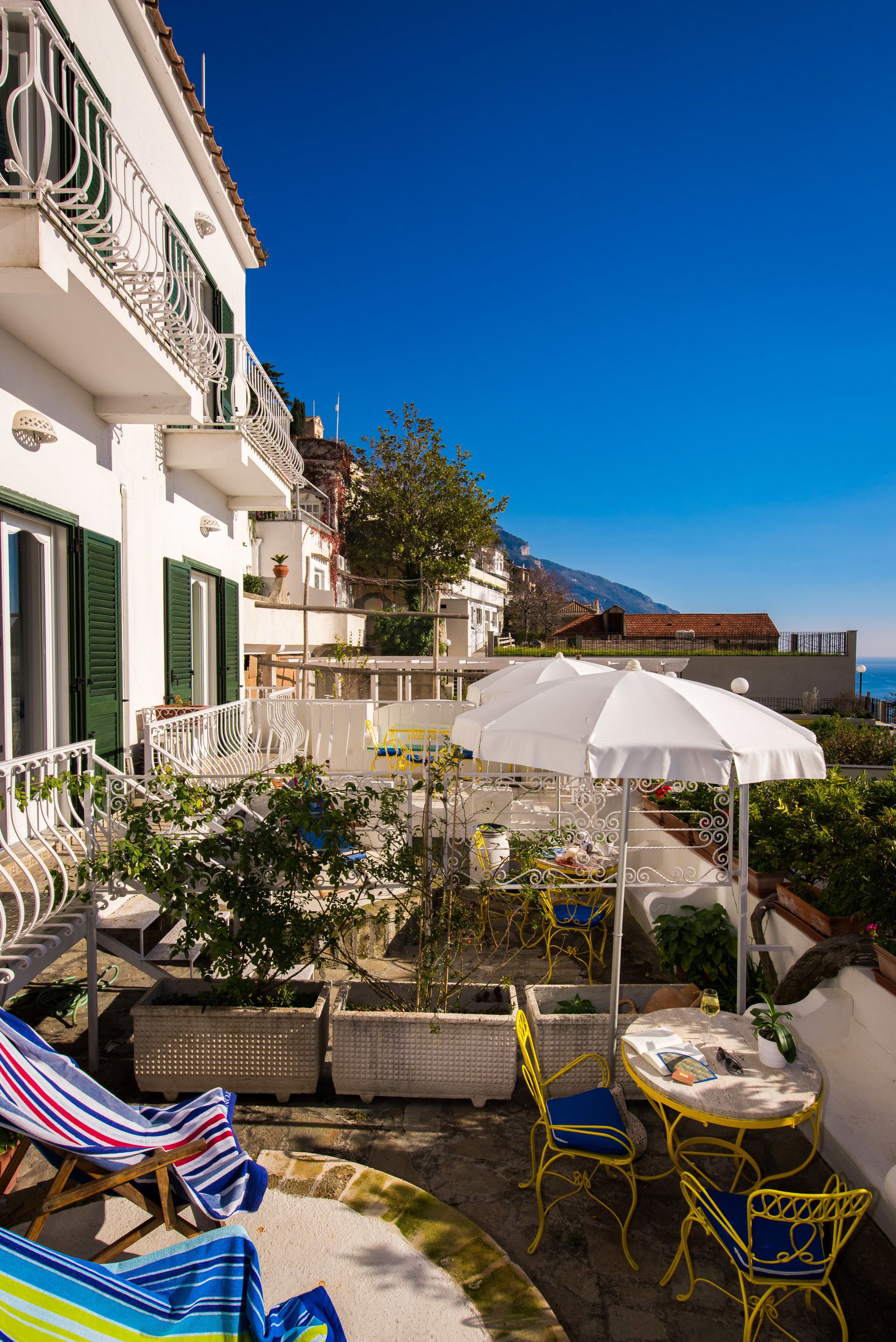 A terrace with outdoor seating, a white umbrella, and a view of the sea and mountains.