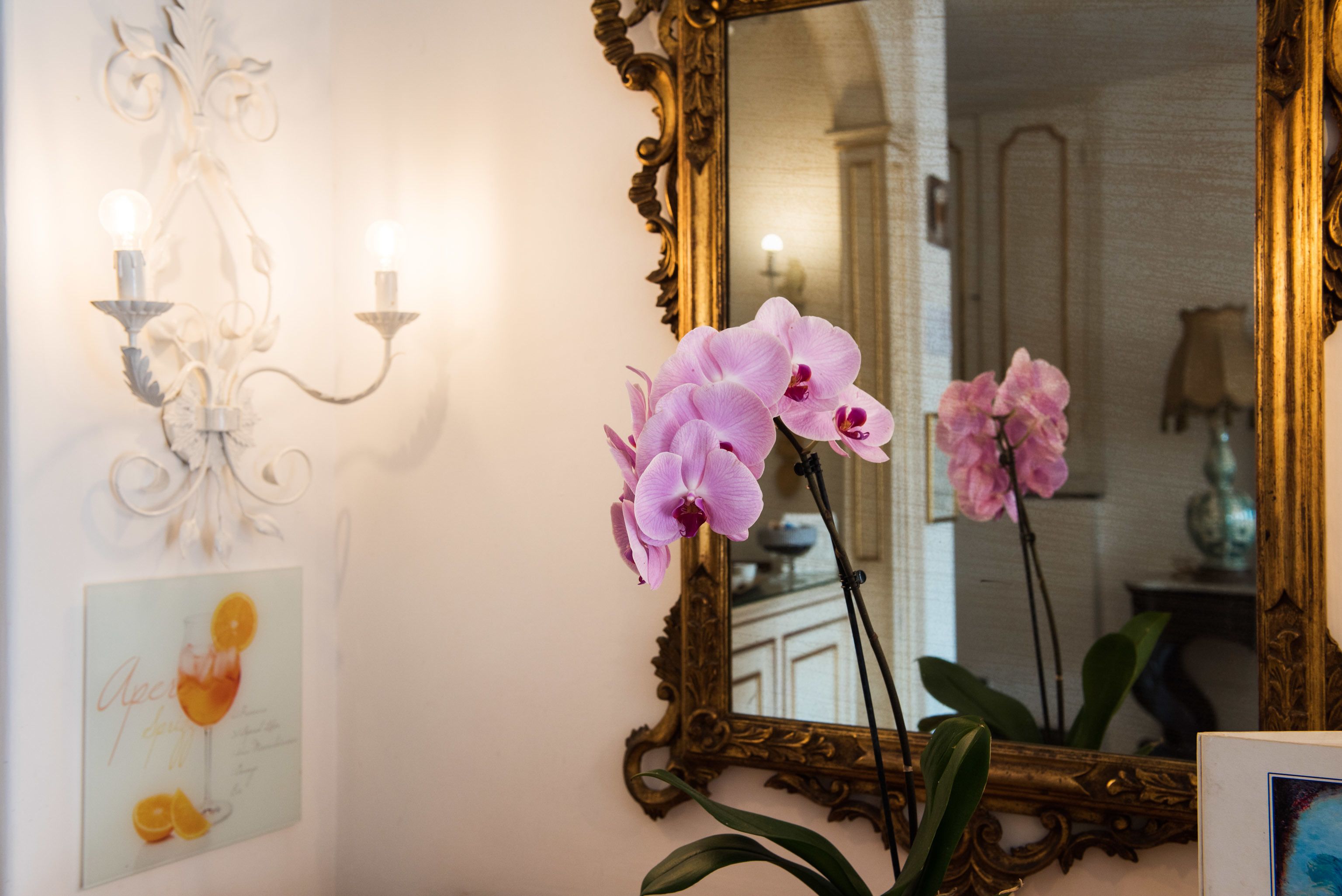 Una stanza con uno specchio grande e decorativo, un candelabro a parete e un quadro che raffigura una bevanda.