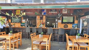 Un café al aire libre con mesas y sillas de madera, una zona de bar con mostrador, y una pantalla de televisión sobre la barra.