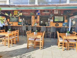 An outdoor cafe with wooden tables and chairs, a bar area with a counter, and a television screen mounted above the bar.