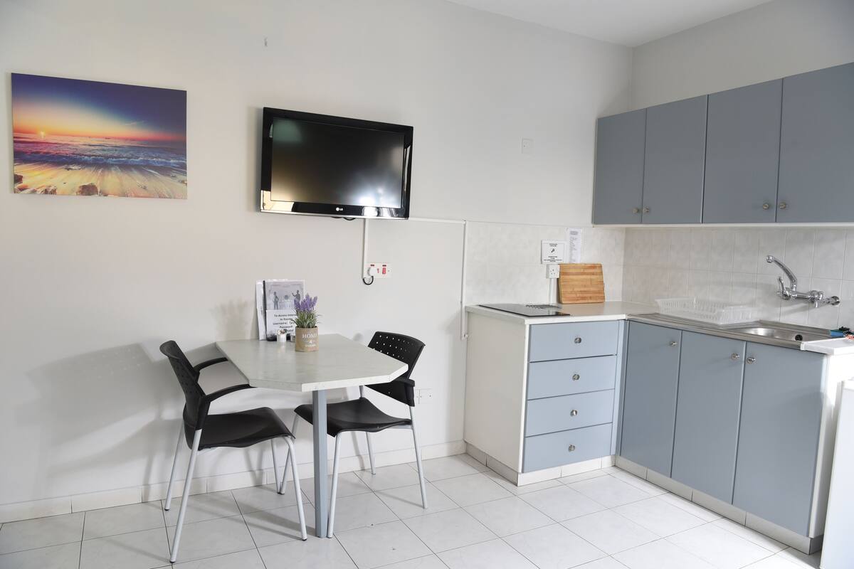 Una cocina moderna con armarios grises, encimera blanca y un lavamanos integrado. Hay una mesa de comedor con sillas y un televisor de pantalla plana montado encima.