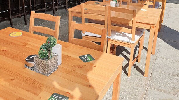 Un área de estar al aire libre con mesas y sillas de madera, una barra con un letrero que dice "TILCOON BAR", y vista de la calle al fondo.