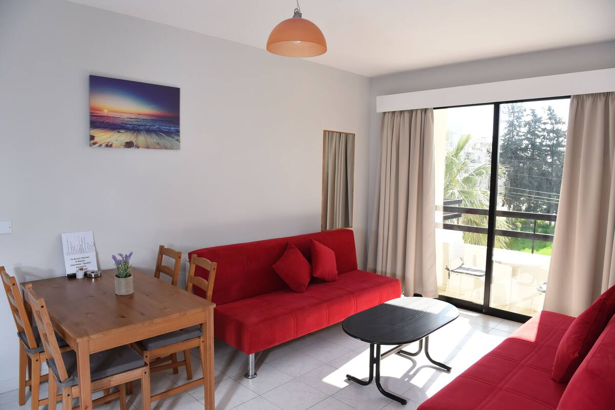 Una sala de estar moderna con un sofá rojo, una mesa de comedor de madera y una mesa de centro negra. Hay un cuadro en la pared y un ventanal con cortinas.