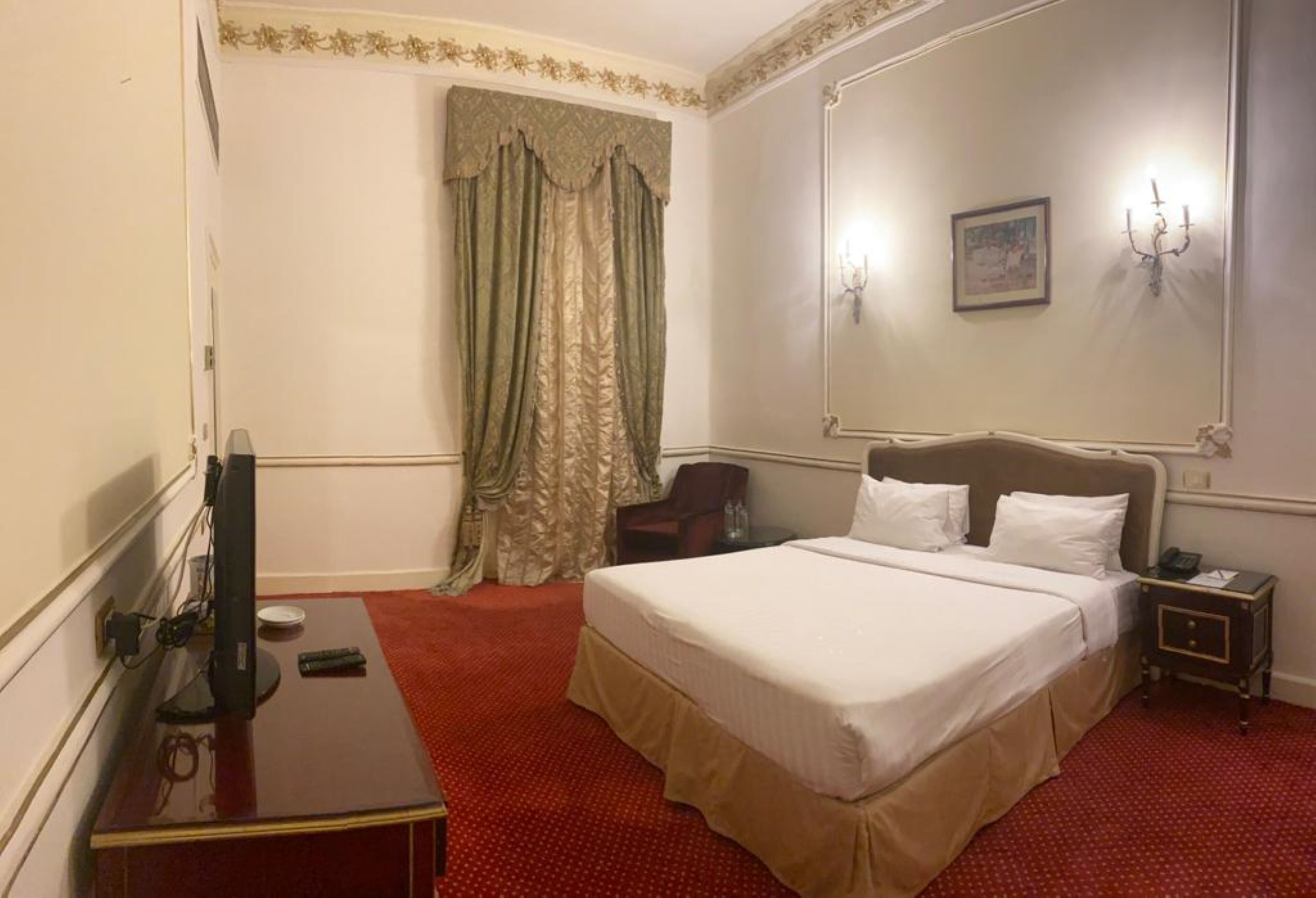 A hotel room with a large bed, a desk with a computer, a chair, a small table, and a nightstand with a lamp.