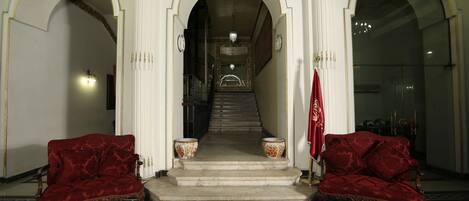 A grand staircase leading to a higher floor, flanked by ornate white columns and red velvet sofas.