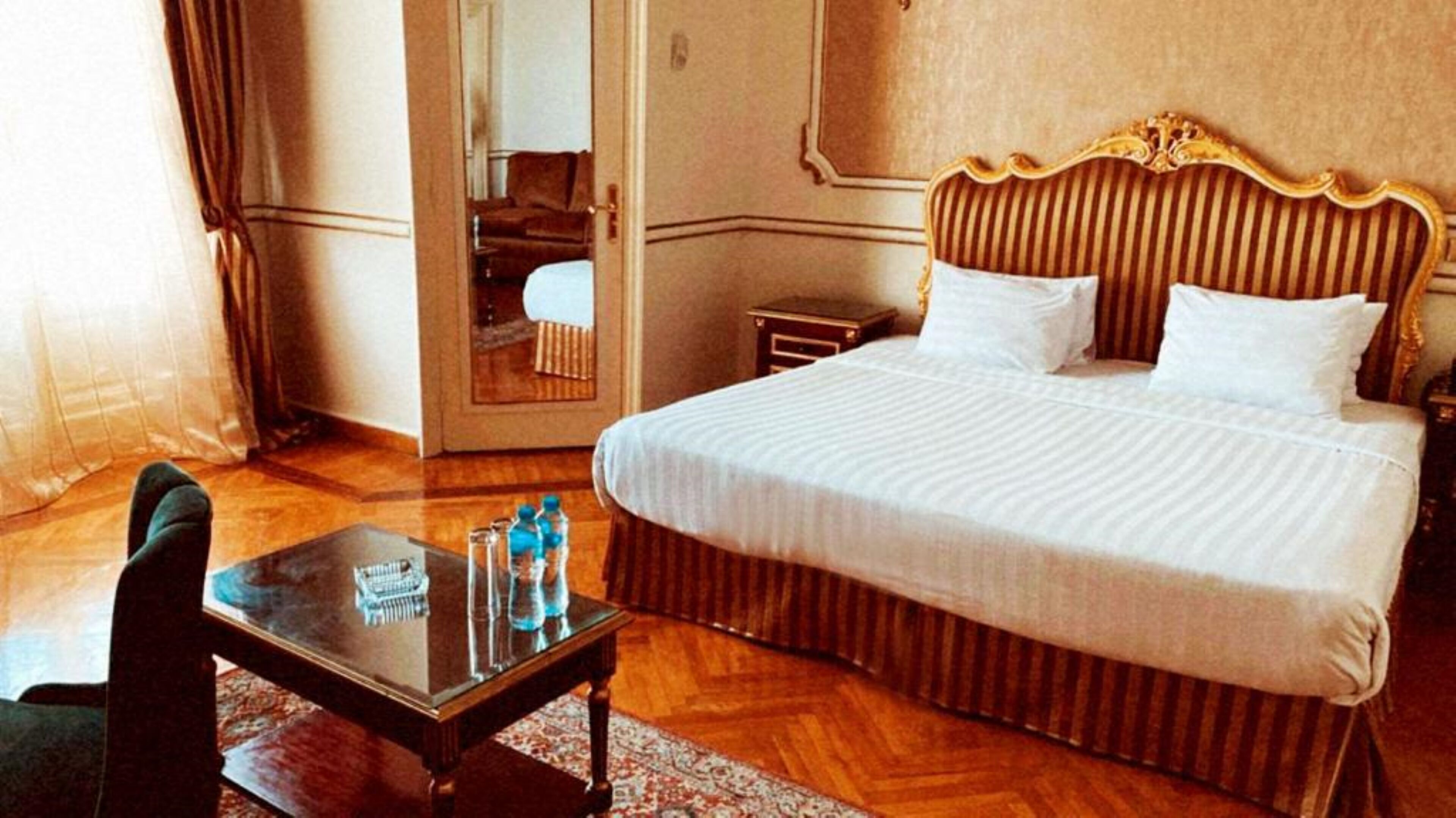 A hotel room with a large bed, a small table with water bottles, a chair, and a mirror.