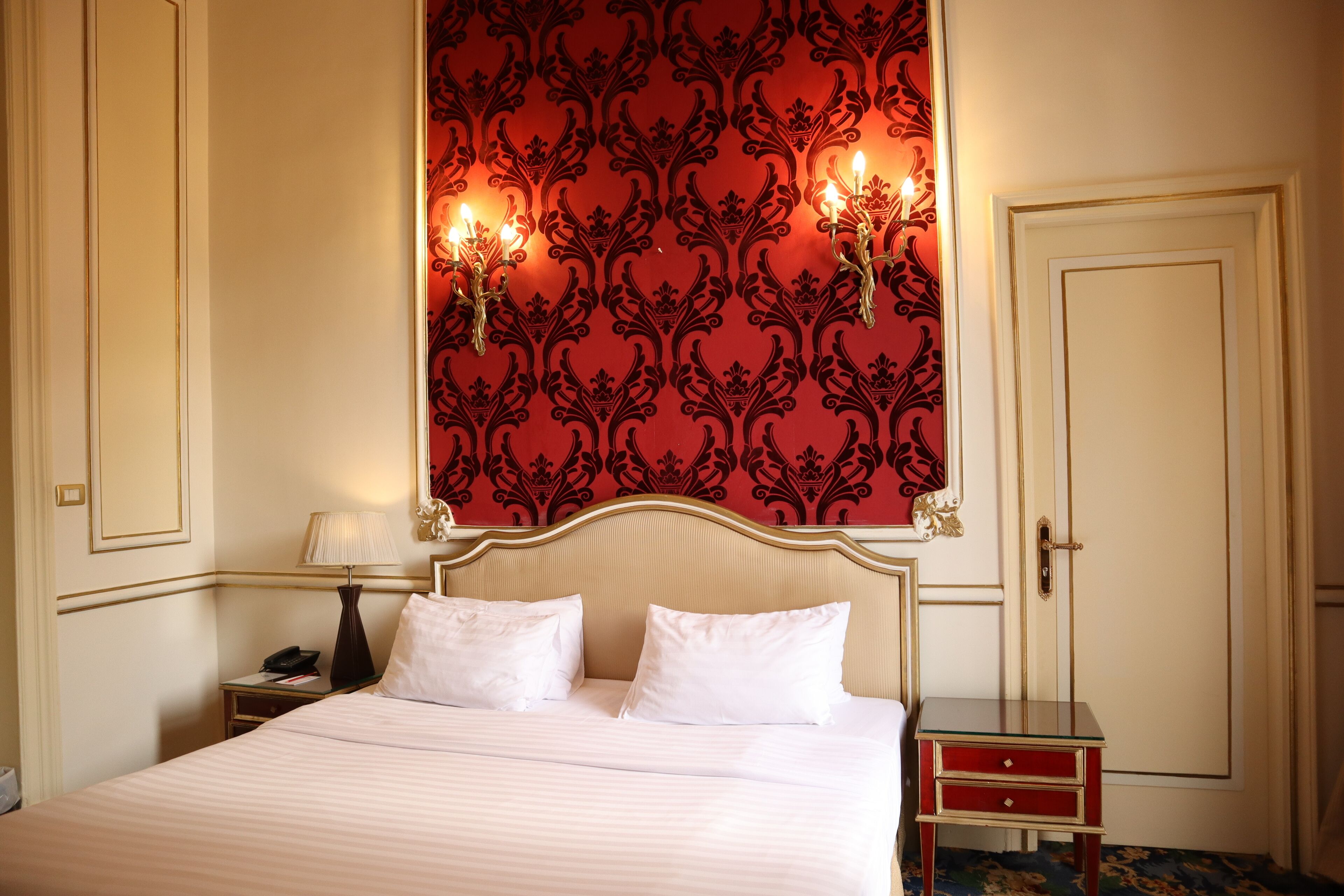 Habitación de hotel con cama, mesitas de noche, lámpara y pared decorada con un papel tapiz estampado en rojo.