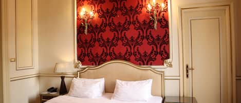 Habitación de hotel con cama, mesitas de noche, lámpara y pared decorada con un papel tapiz estampado en rojo.