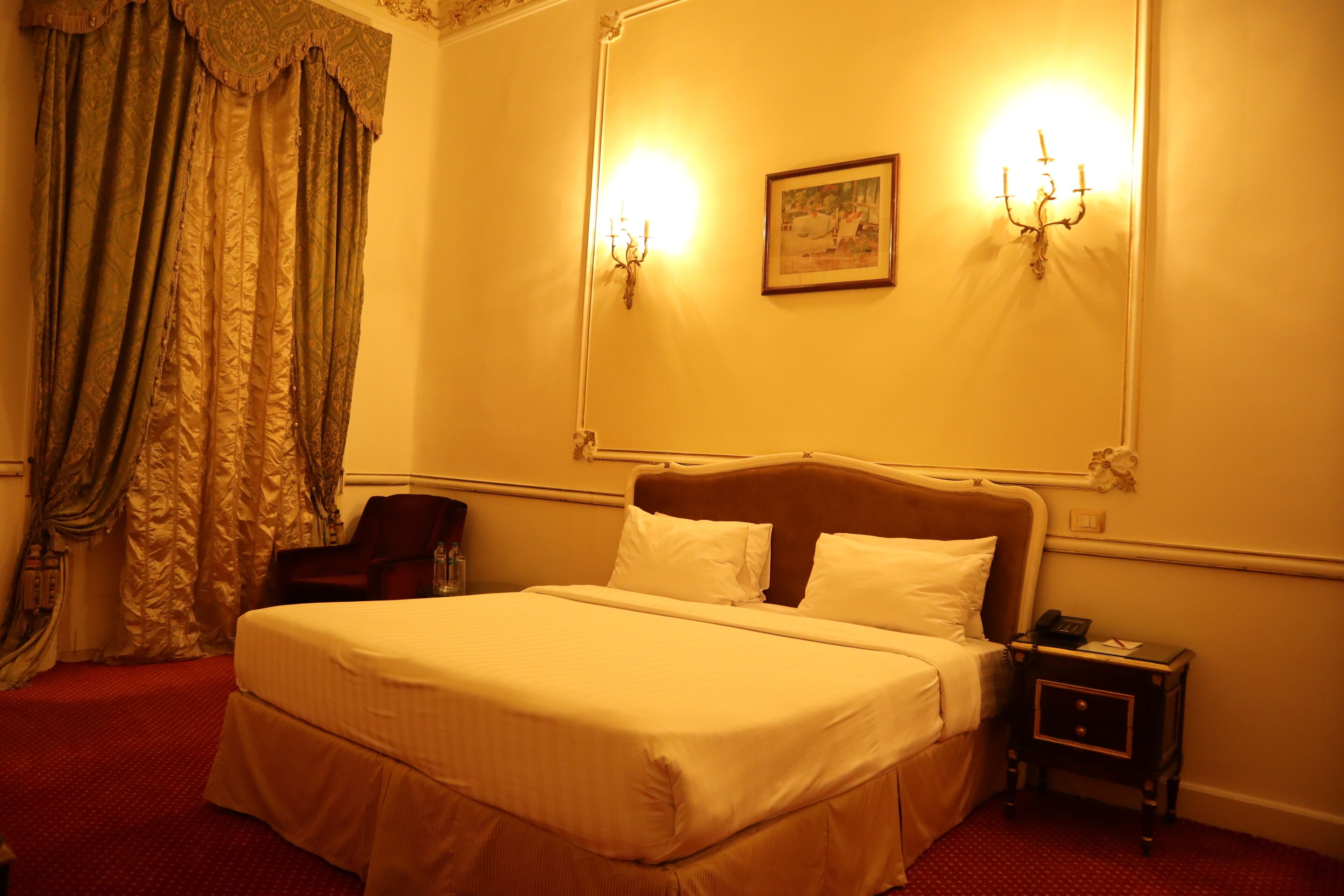 A hotel room with a large bed, a nightstand, a chair, and a painting on the wall.