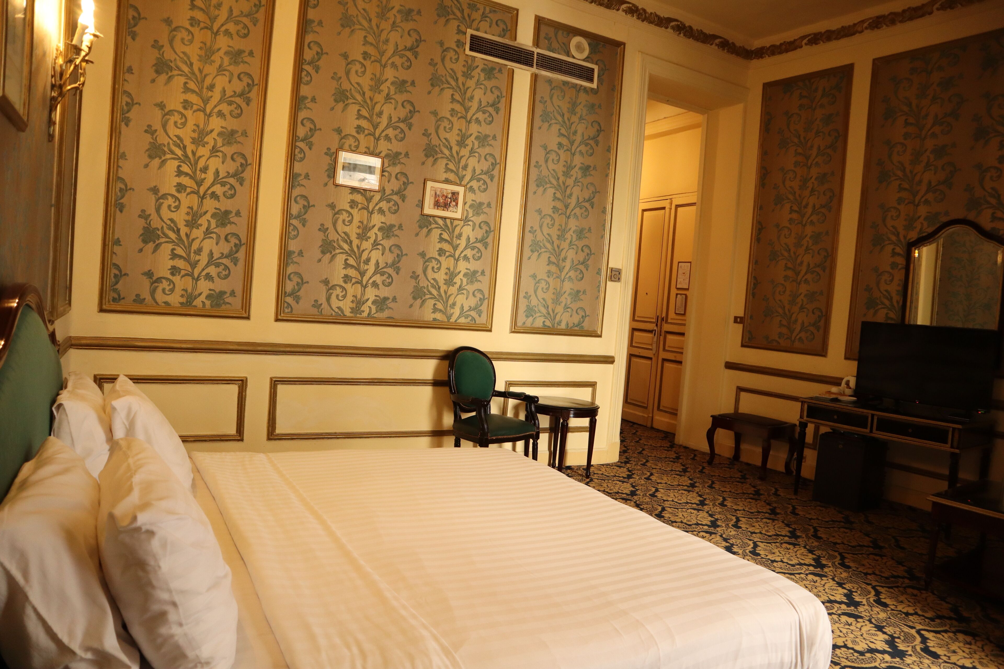 A hotel room with a large bed, a desk, a chair, a television, and a wardrobe.