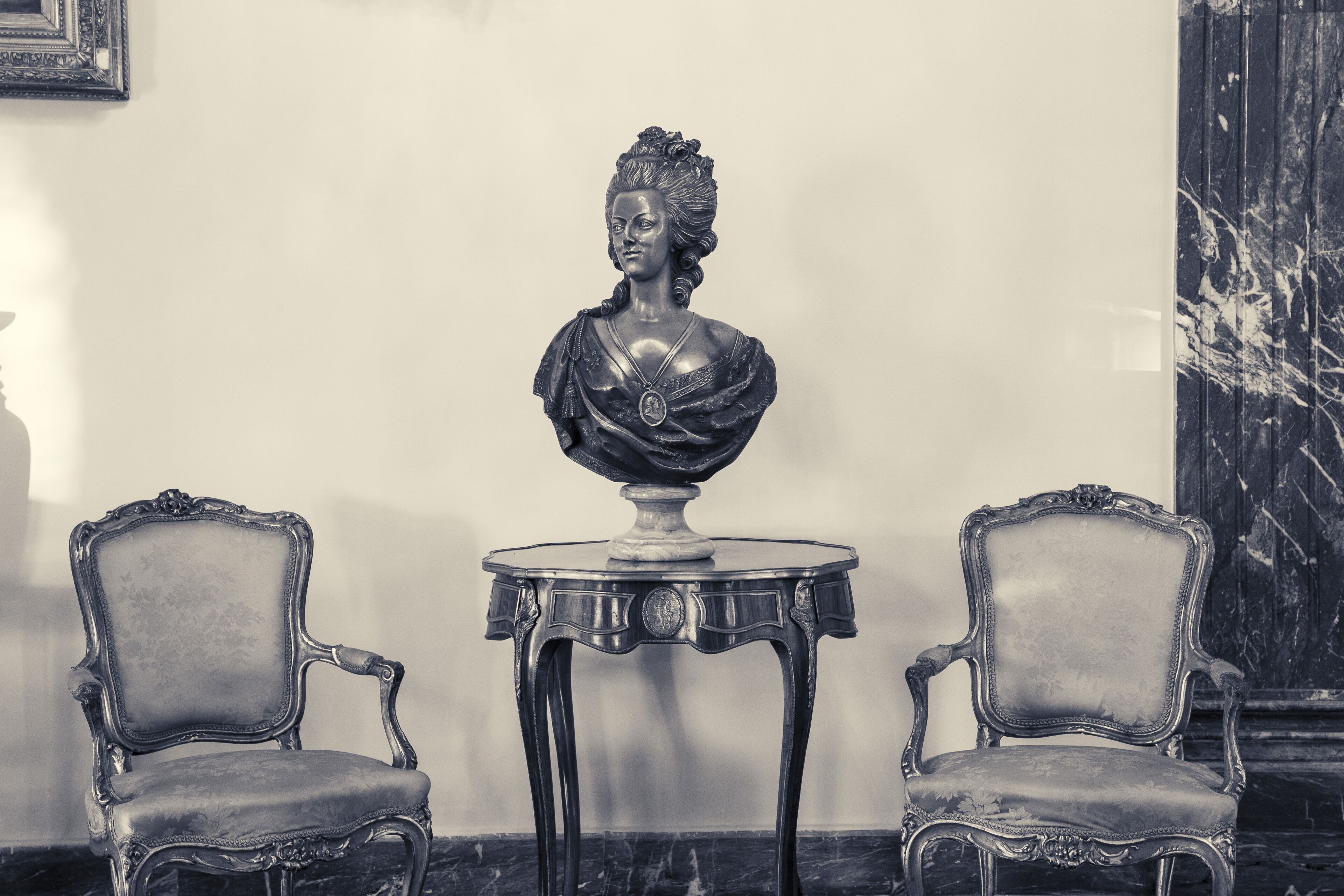 A room with a bust on a pedestal table, flanked by two ornate chairs.
