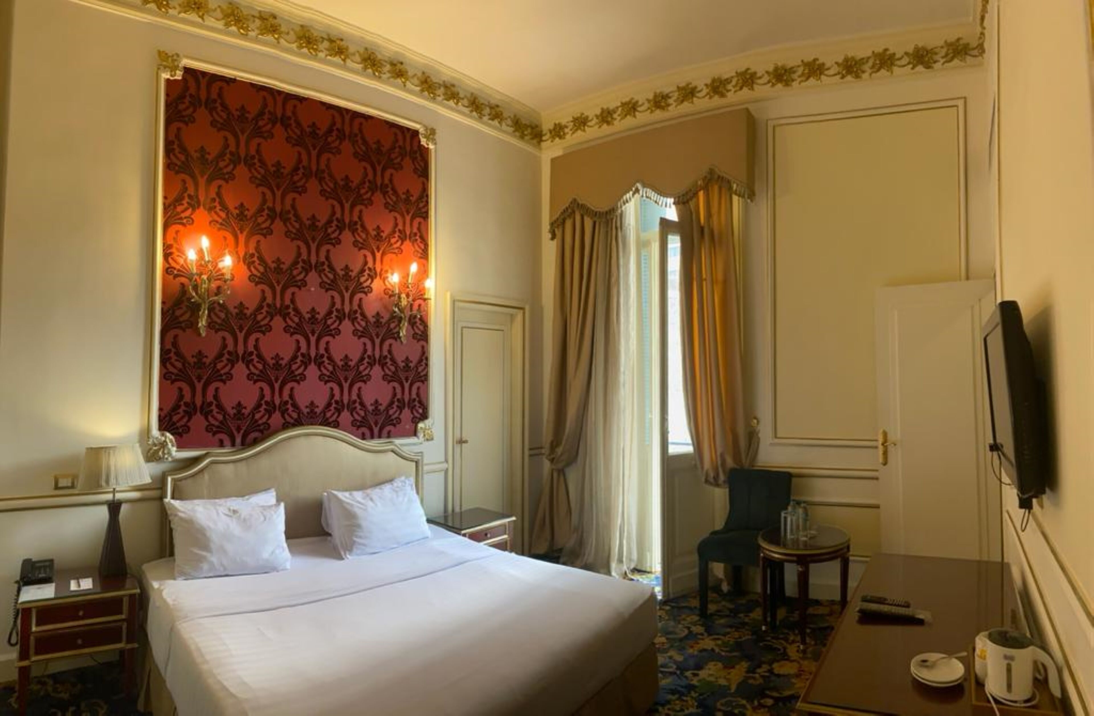 Une chambre d’hôtel avec un grand lit, un bureau avec une lampe, une chaise, une petite table avec une tasse et une soucoupe, et un téléviseur fixé au mur.