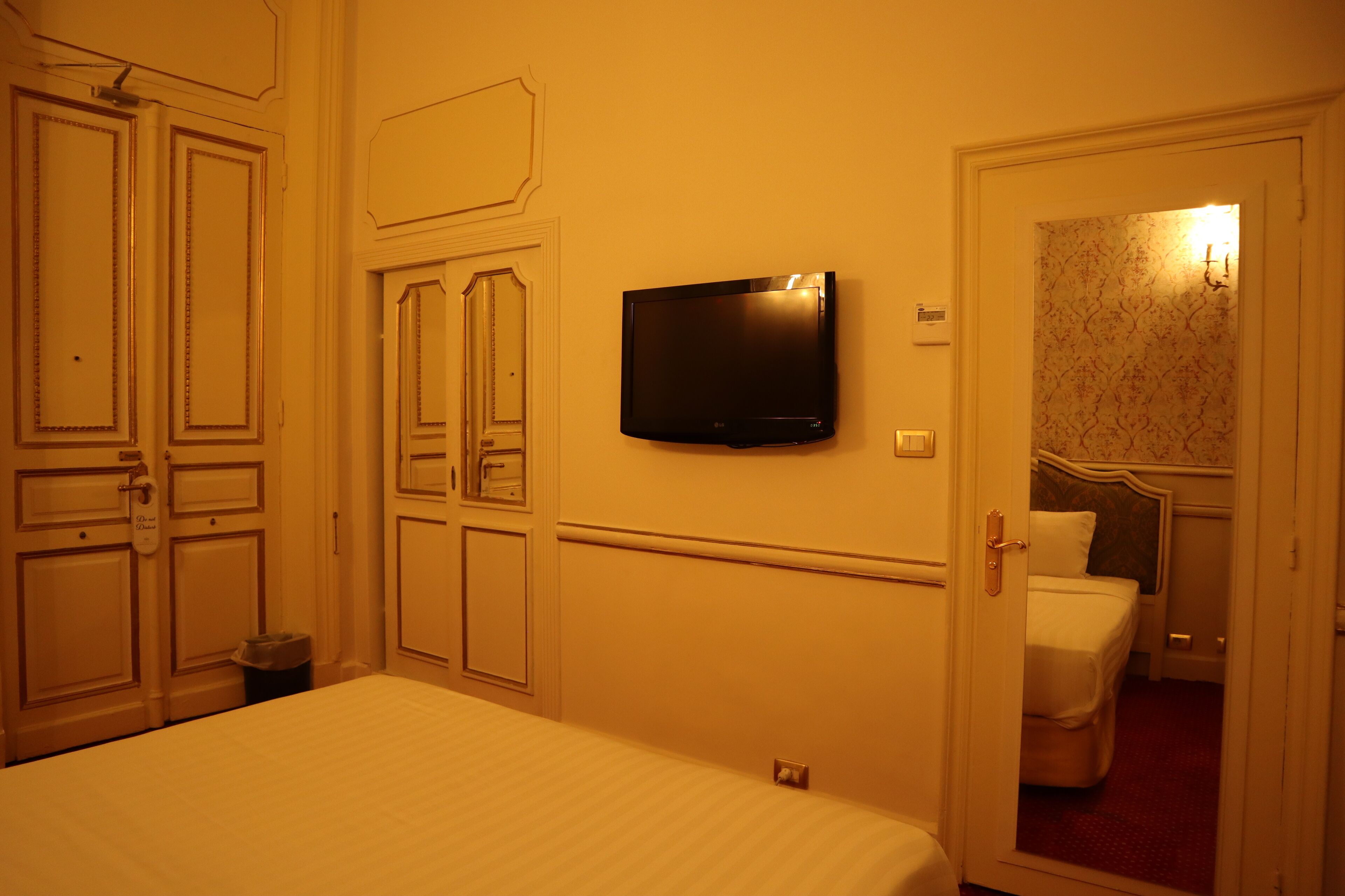 Quarto de hotel com uma cama, uma televisão fixada na parede e uma porta que dá para outro espaço.