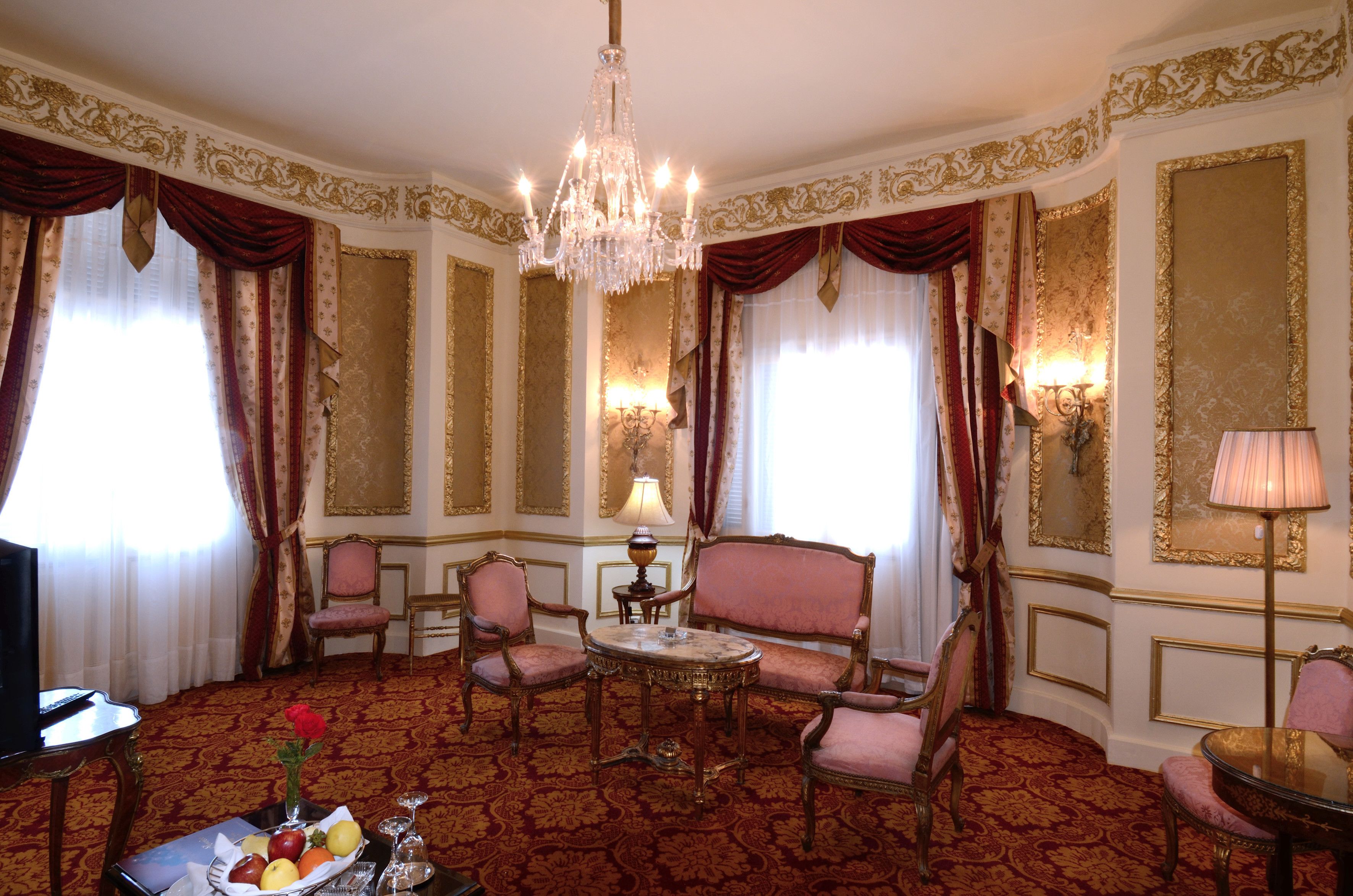 Une chambre luxueuse, ornée d’un lustre, de fauteuils à revêtement rouge, d’une table ronde et d’un téléviseur.