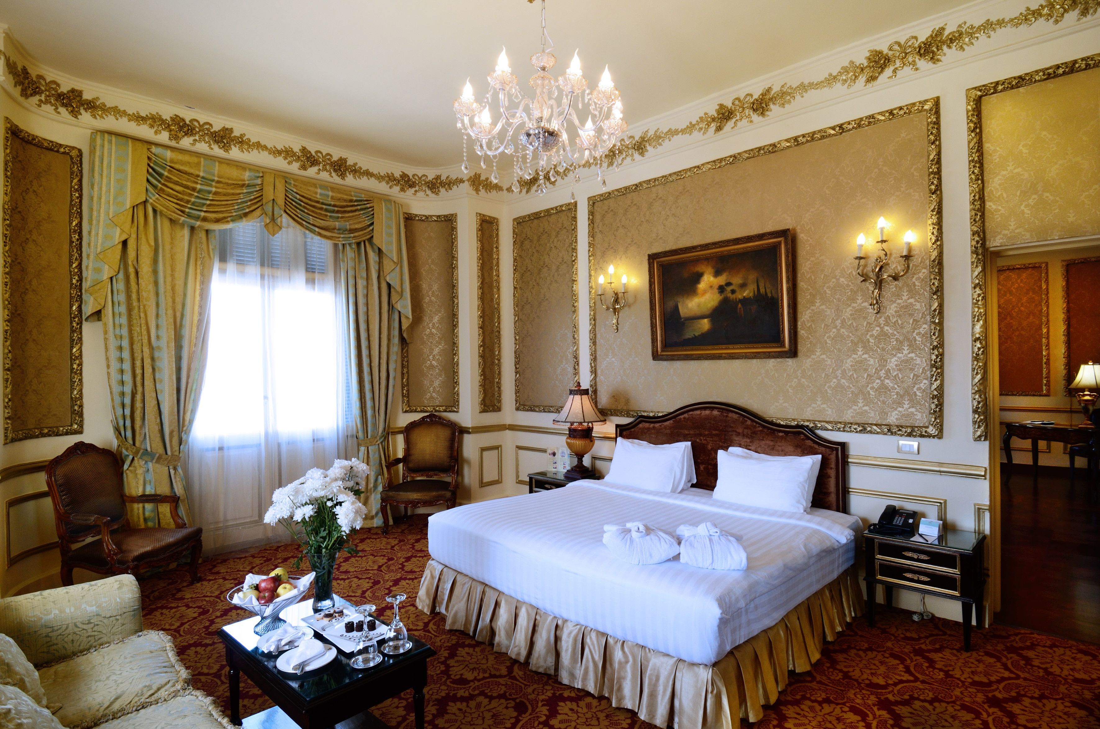 A luxurious hotel room with a large bed, a chandelier, a painting on the wall, and a desk with a lamp.