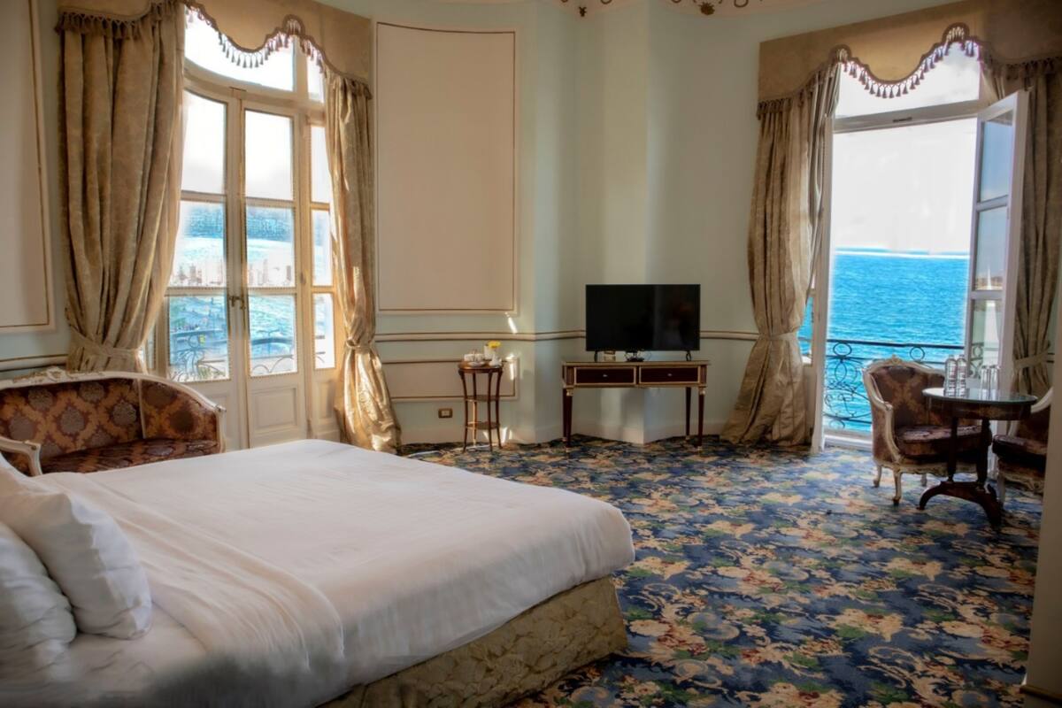 Habitación de hotel con una cama grande, televisión, una mesita y una silla. Cortinas y vistas al mar a través de las ventanas.