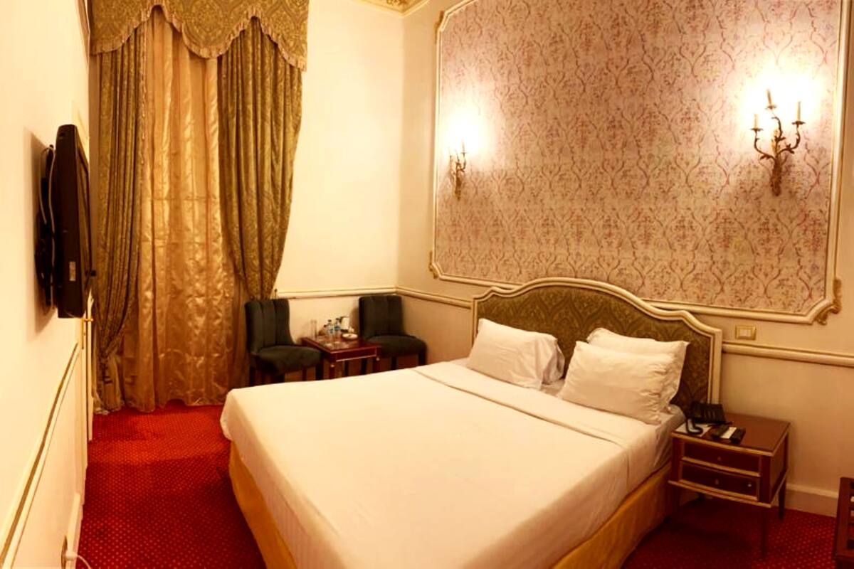 Una habitación de hotel con una cama grande, dos sillas, una mesita de noche y una lámpara de pared.
