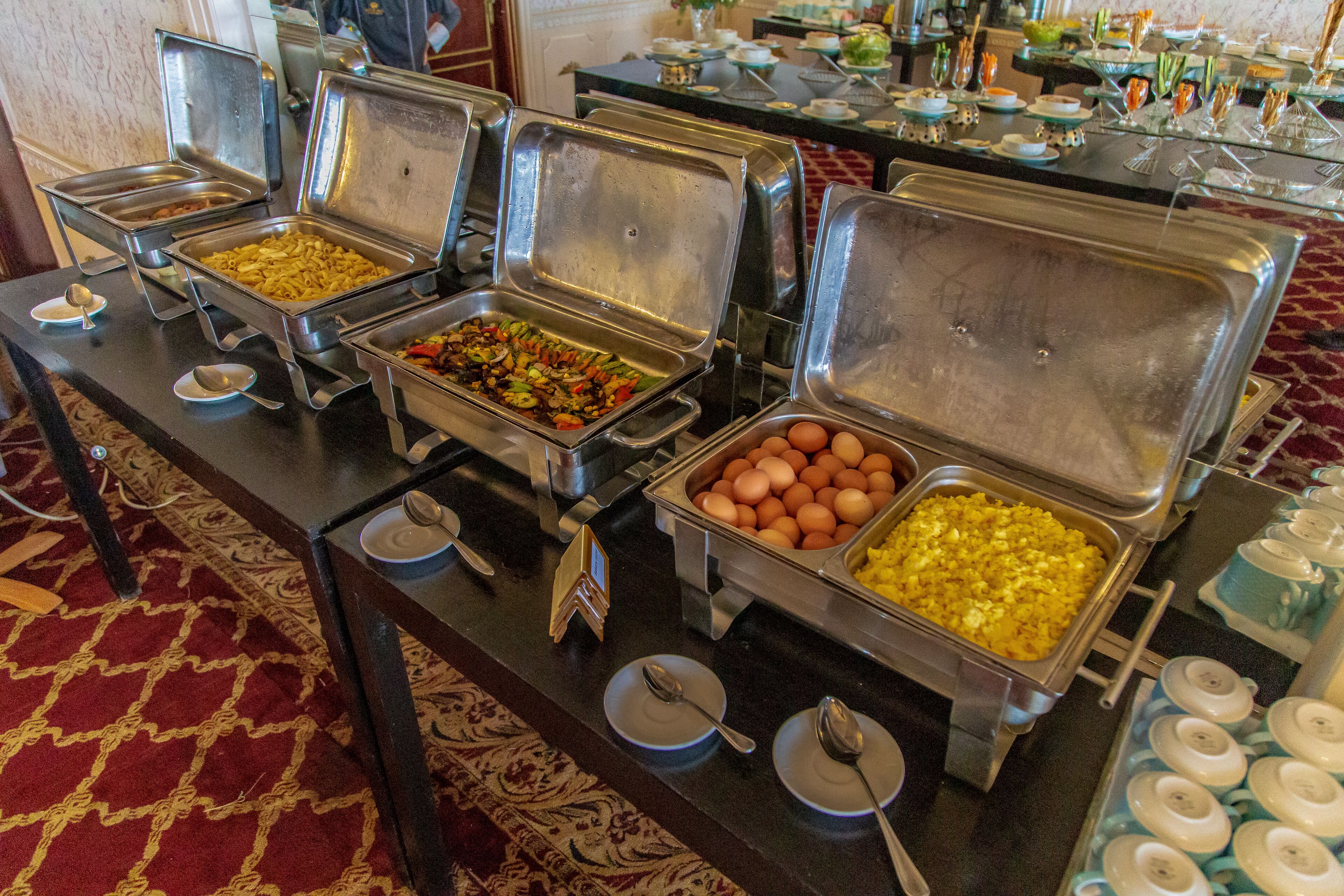A buffet setup with various food items in metal containers, arranged on a table with a patterned carpet.