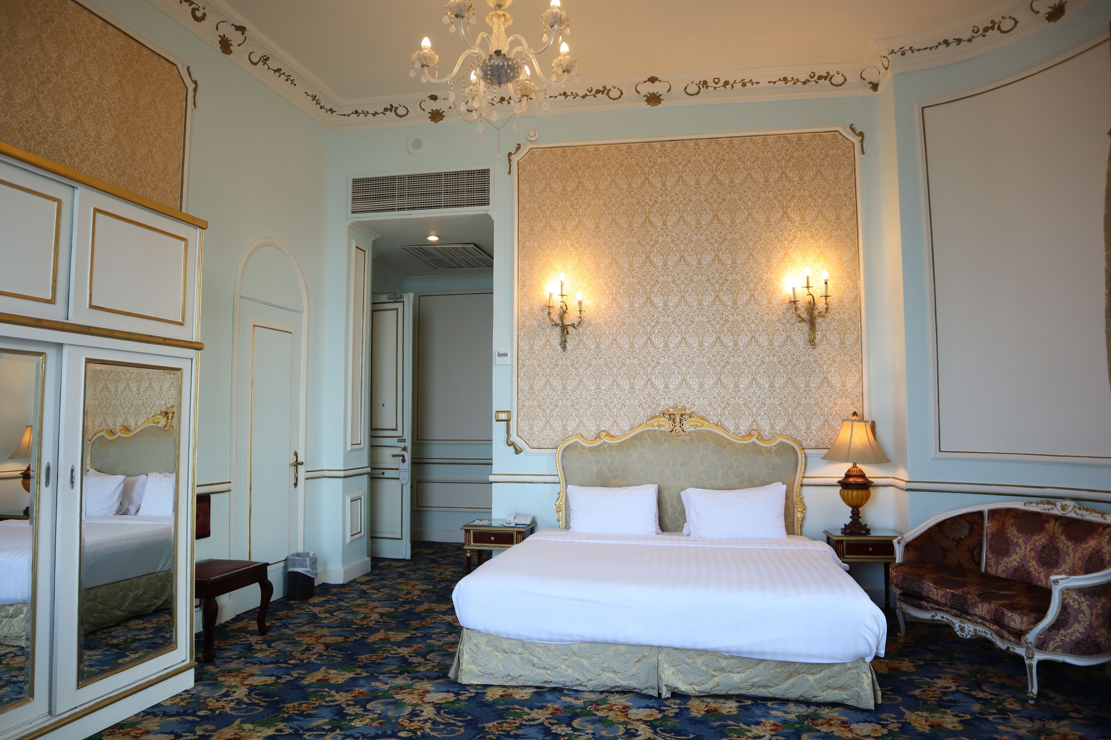 A hotel room with a large bed, a mirror, a chandelier, and a patterned carpet.