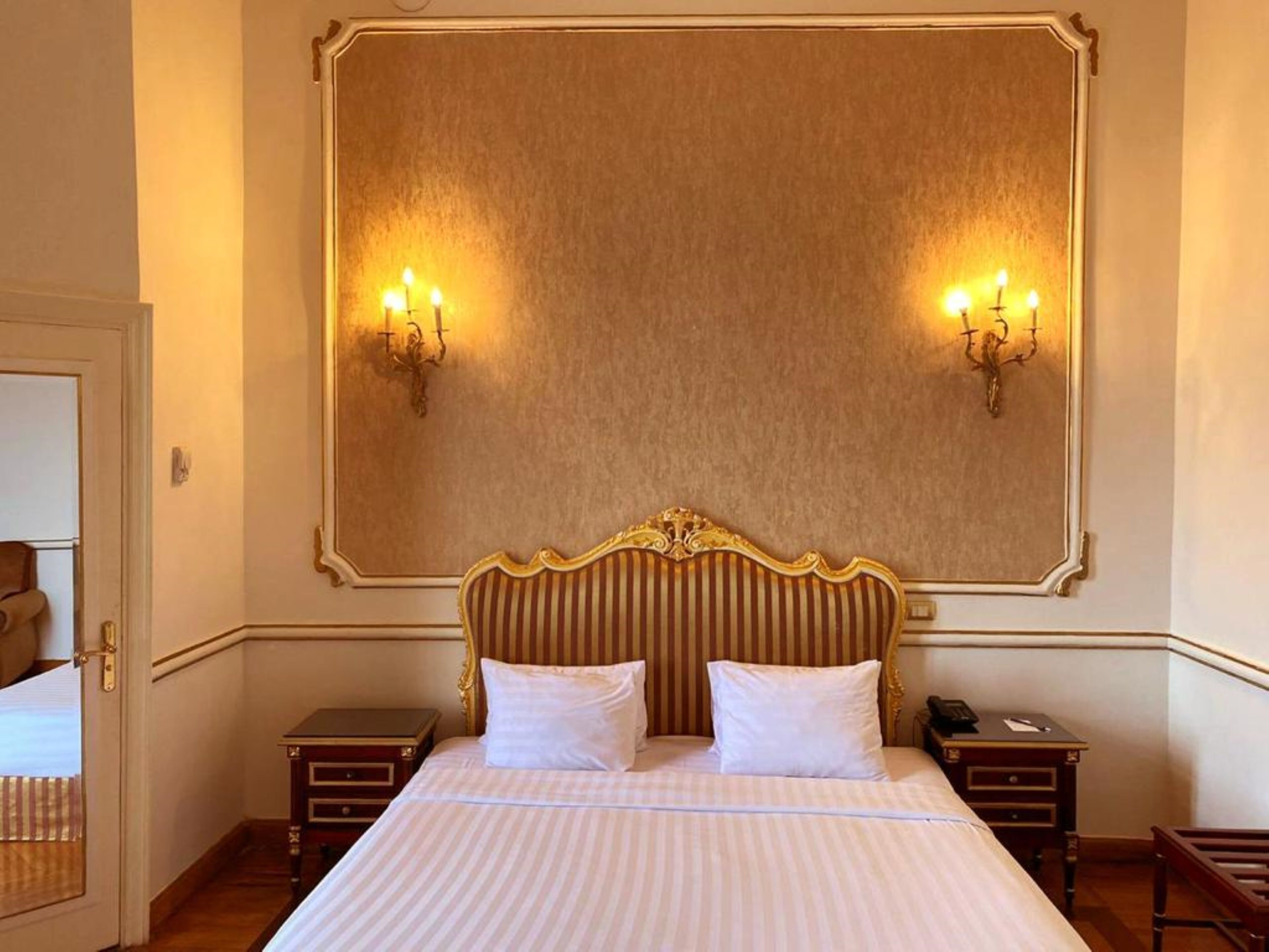A hotel room with a large bed, two bedside tables, a mirror above the bed, and wall sconces.